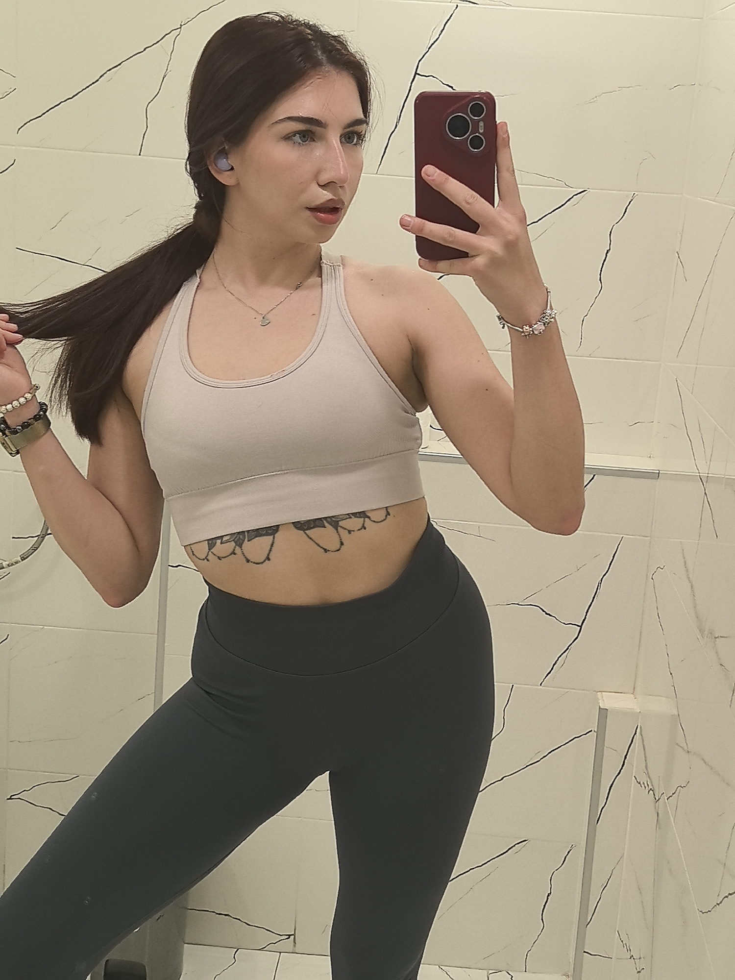 A young woman is taking a selfie in what appears to be a bathroom. The walls are covered in white marble-like tiles with black veining.
She is wearing a light-colored sports bra and dark leggings, exposing a tattoo across her lower abdomen. She is holding a red phone in her right hand to take the photo. She has long, dark hair pulled back into a ponytail and is wearing bracelets on both wrists and a necklace. She also seems to have an earbud in her left ear.
The location cannot be determined to be Stara Zagora, Bulgaria from the image alone. The background only reveals the bathroom interior.