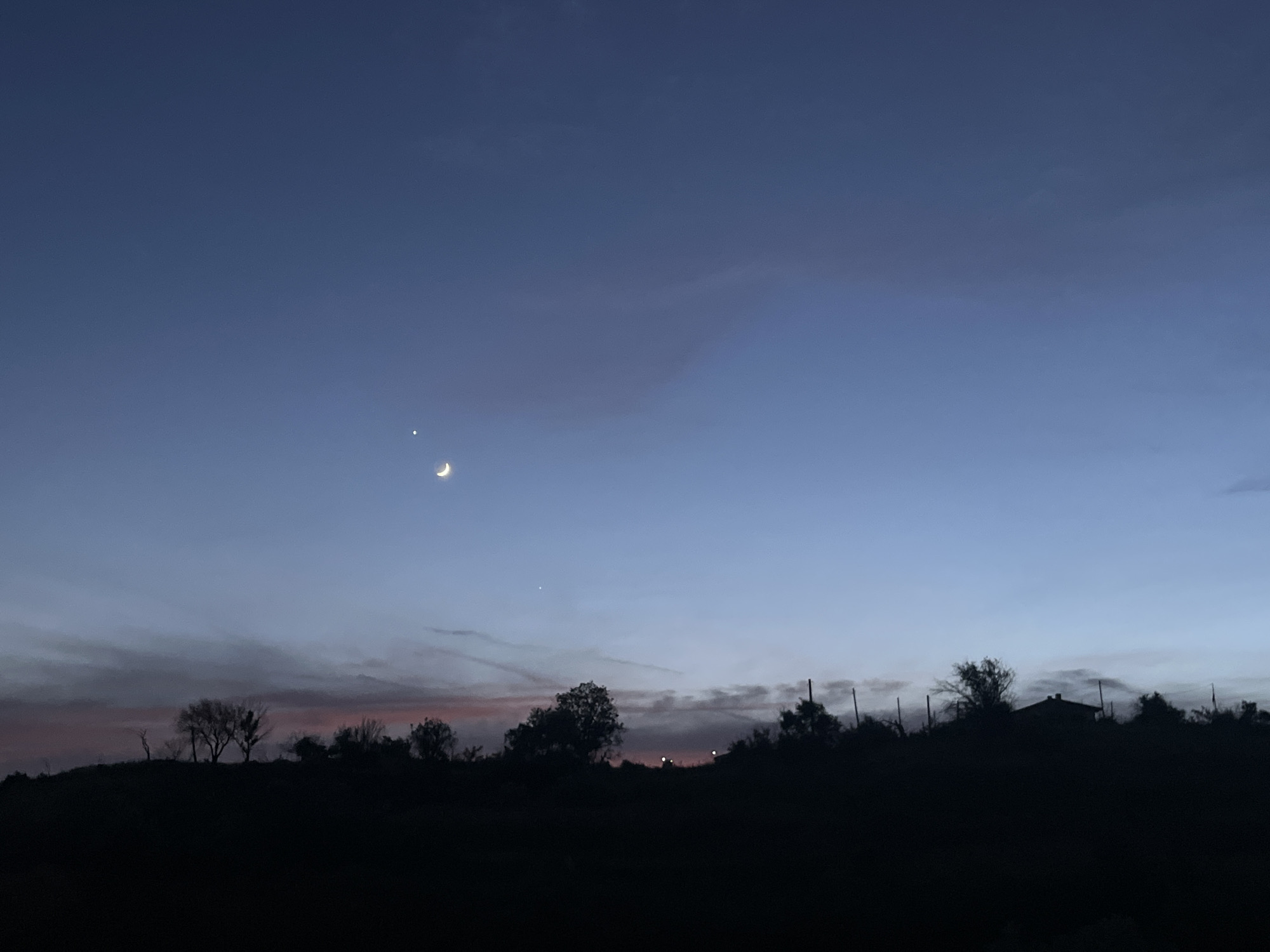 The image depicts a twilight scene with a dusky blue sky transitioning to lighter hues near the horizon. A crescent moon and a bright star, likely Venus, are visible in the upper portion of the sky. The foreground is dominated by a dark silhouette of a hilly landscape, featuring trees and the outline of a building. The scene suggests a rural or suburban setting, possibly on the outskirts of Varna, Bulgaria, given the context. There are no people or specific actions depicted. The atmosphere is serene and tranquil, characteristic of the transition from day to night.