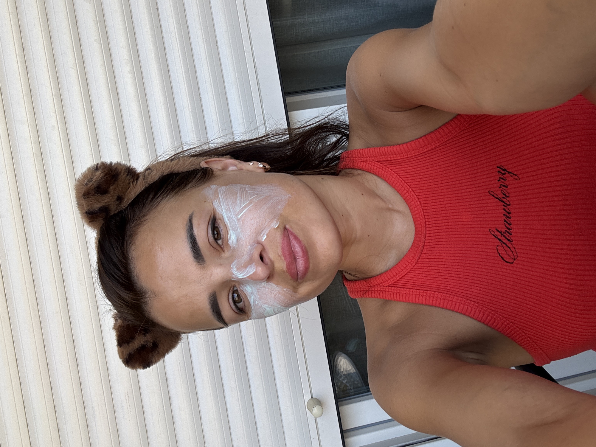 A young woman with brown hair is wearing a red tank top and a brown fuzzy headband with bear ears. She has a white mask on her face and is smiling. She is outdoors in front of a white, slatted door.