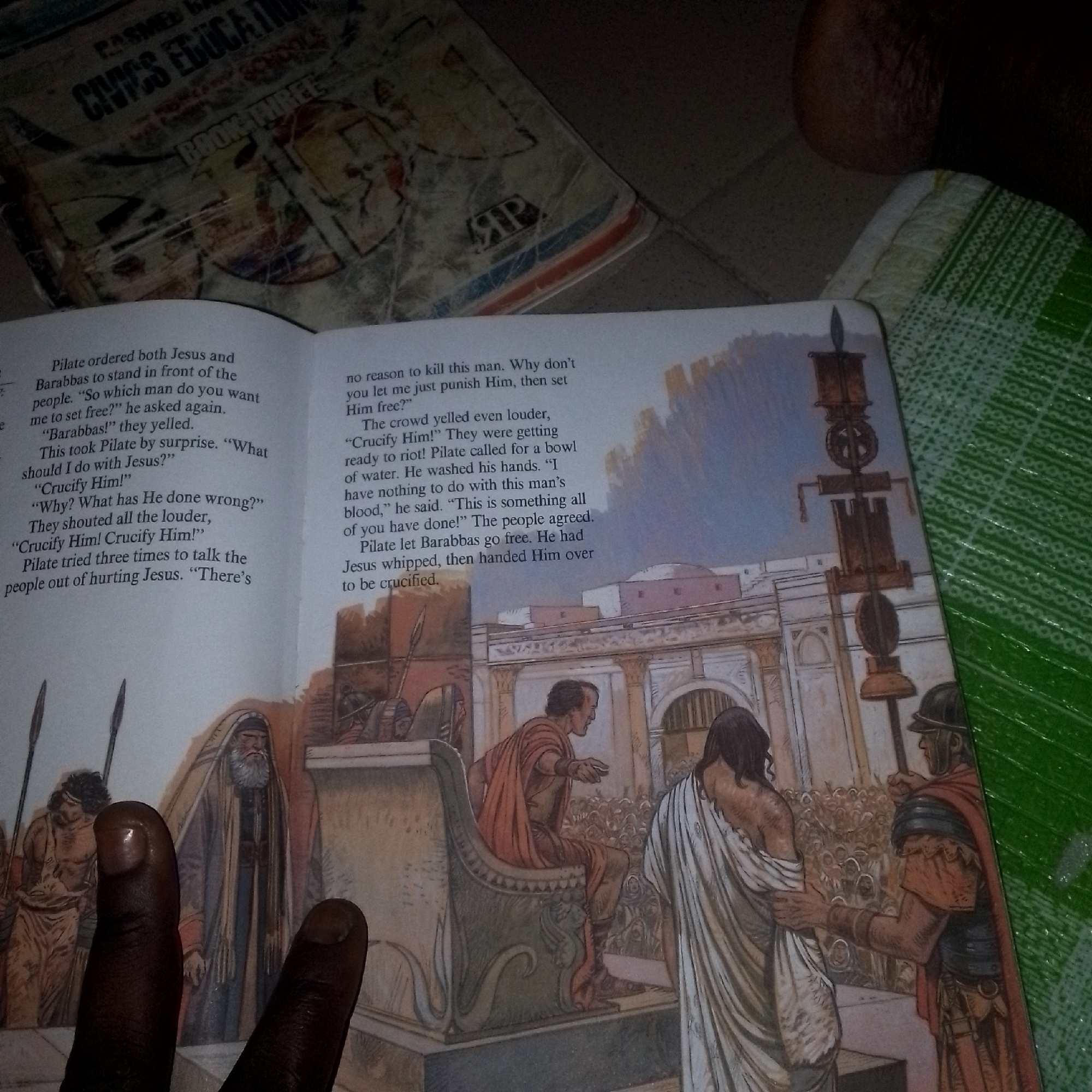In a scene depicted on an open book, a passage describes a historical event. The text details Pilate's decision regarding Jesus and Barabbas. In the background, an illustration shows a Roman-style architecture with columns and arches. Several figures are present, including a man seated on a throne-like structure, a figure draped in white, and a man in armor holding a staff. Two hands are visible in the foreground, obscuring a portion of the illustration. The book is resting on a green textured surface. The top of the image shows a partially obscured book cover with text that appears to read "CIVICS EDUCATION".