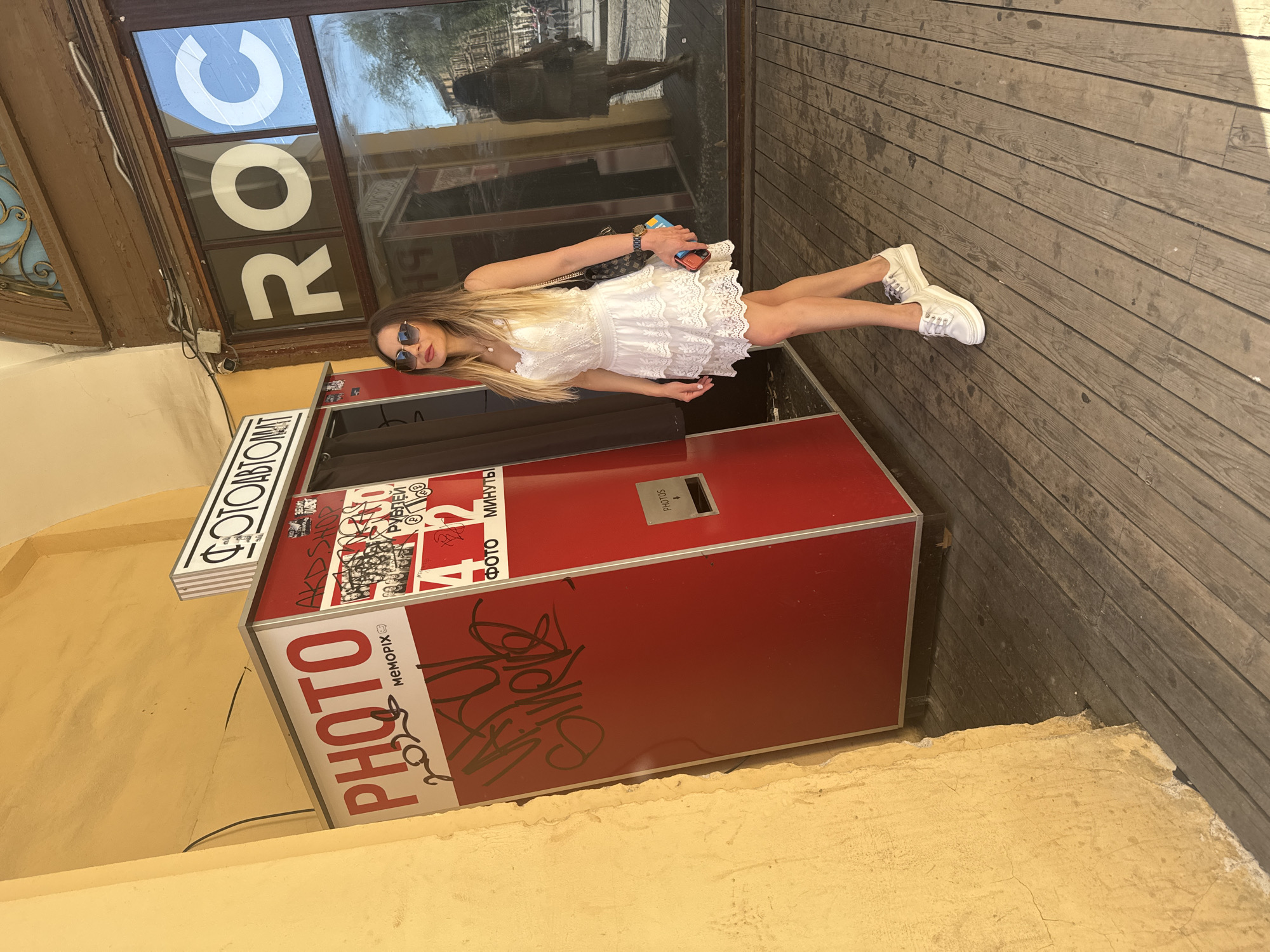 A young woman stands in front of a red photo booth in an outdoor setting. She is wearing a white lace dress and white sneakers, and sunglasses. She holds a small purse and a wallet or phone in her hand. The photo booth has "PHOTO" written on it in large red letters, and there are other signs and graffiti on it. In the background, a wooden floor and a building with a large window displaying the word "ROC" are visible. The scene appears to be taking place during the daytime, under bright sunlight. The woman has a relaxed posture, suggesting a casual outing. The graffiti and wear on the photo booth suggest it might be in a public space or tourist area. The word "Saint Petersburg" in the location context implies the woman is in Russia.