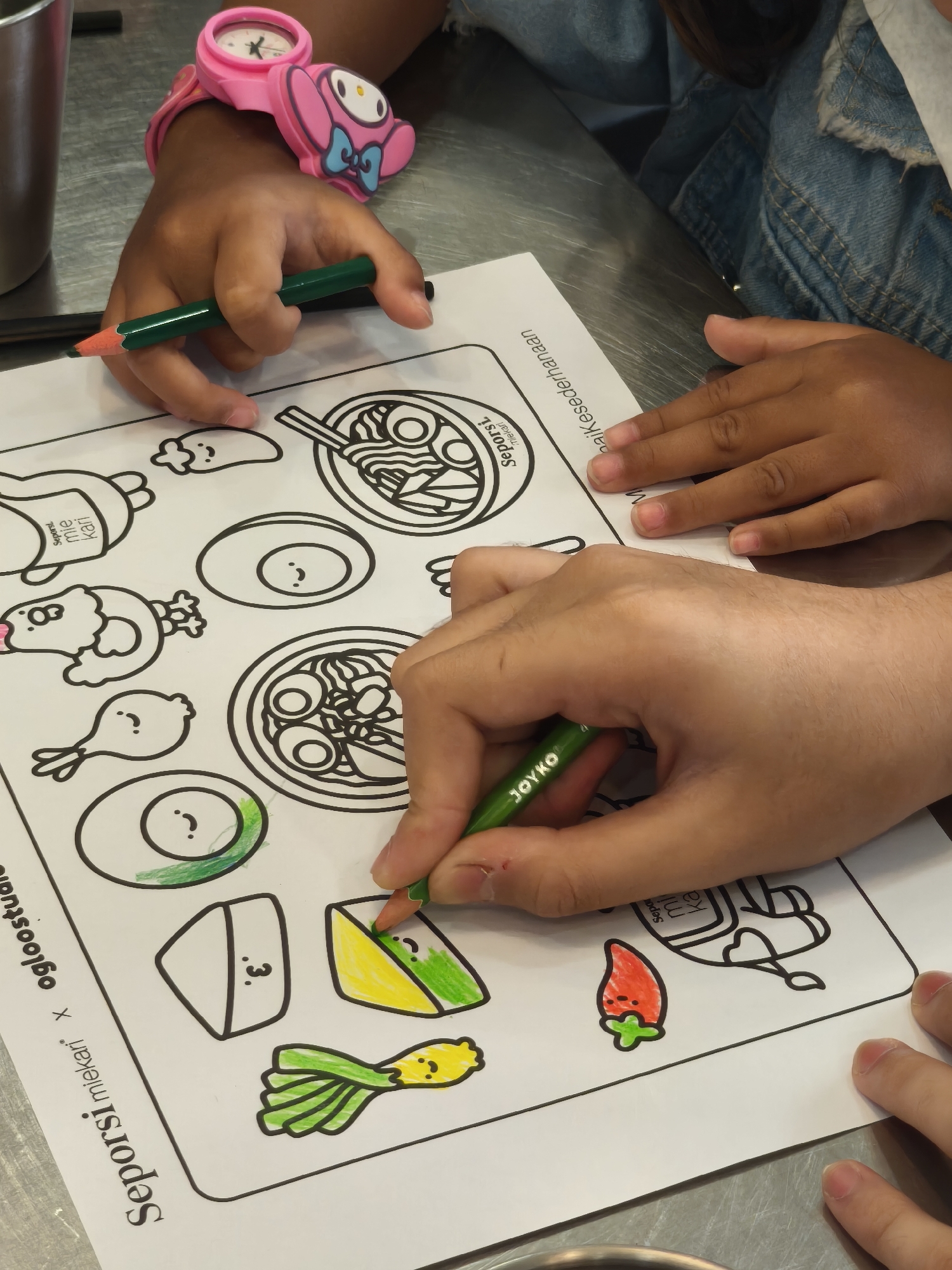 In South Jakarta, Indonesia, an overhead shot captures two sets of hands coloring a printed sheet featuring various cartoon food items. The left hand, belonging to a child wearing a pink Hello Kitty watch, holds a green crayon and is poised to color. The right hand, belonging to an adult, grips a green crayon labeled "JOYKO" and is actively coloring a section of a food drawing. The coloring sheet is positioned on a metallic surface, suggesting a table.