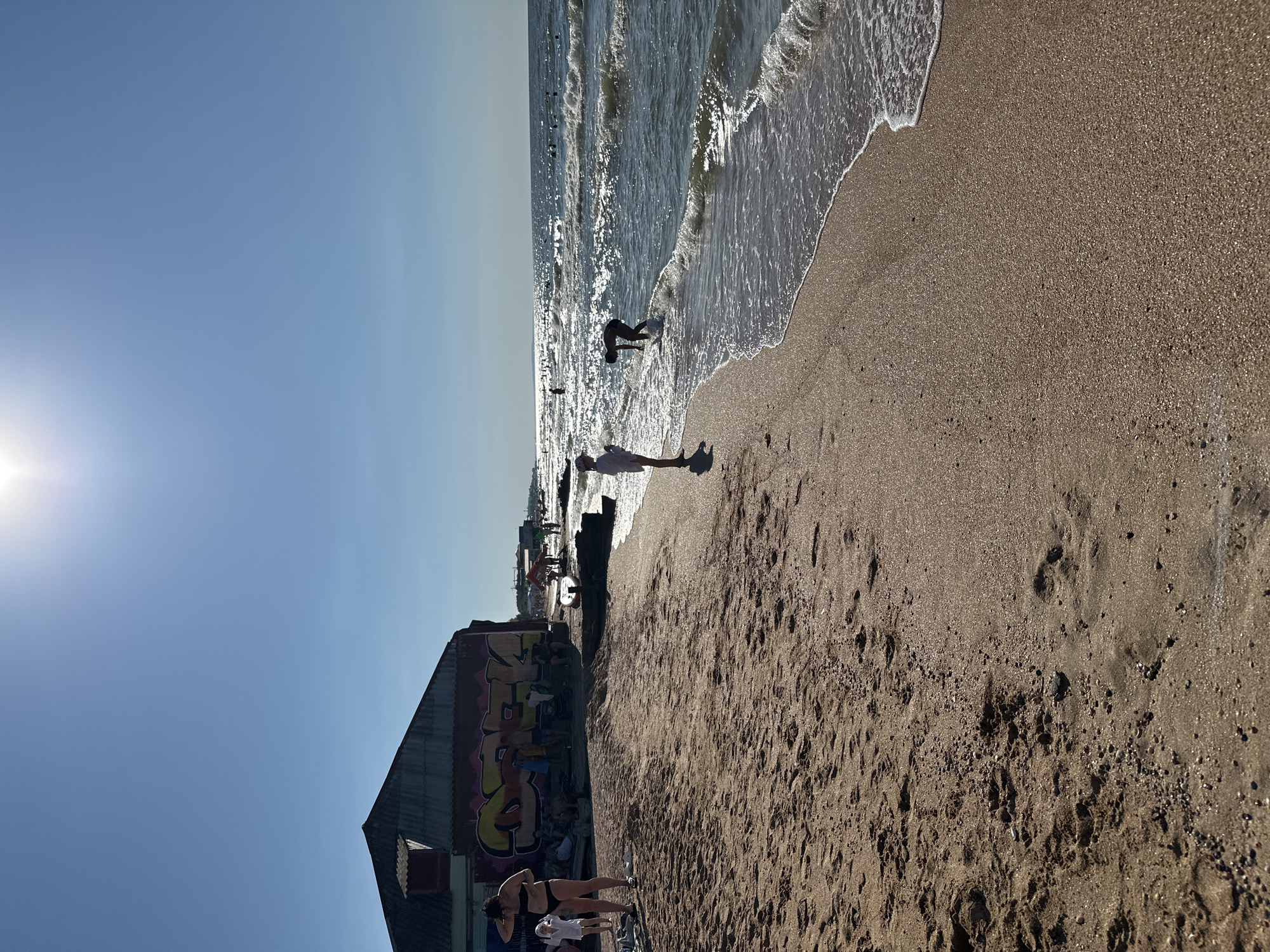 This image depicts a sunny day at a beach in Golubitskaya, Russia. The scene is dominated by a clear blue sky and the sparkling waters of the sea, with gentle waves lapping onto the sandy shore. In the foreground, the sandy beach shows footprints and small pebbles.
Several people are present. Near the water's edge, a person is seen bending down, seemingly interacting with the waves. Further up the beach, a person in a white top and dark shorts stands facing away from the camera, their shadow cast on the sand. Closer to the left of the frame, a building with a corrugated roof is covered in colorful graffiti, with several individuals gathered around its base. Two women in bikinis are visible in this area, one in a black bikini and the other in a white outfit, with one looking down and the other appearing to adjust their hair.
Other people can be seen in the distance along the shoreline and in the water, suggesting a leisurely day at the beach. The bright sunlight indicates it is likely midday or early afternoon. The overall mood appears relaxed and summery, with people enjoying a day outdoors by the sea. The graffiti on the building is a notable detail, adding a touch of urban art to the natural beach setting. There is no visible text other than the graffiti itself, which is difficult to decipher.
