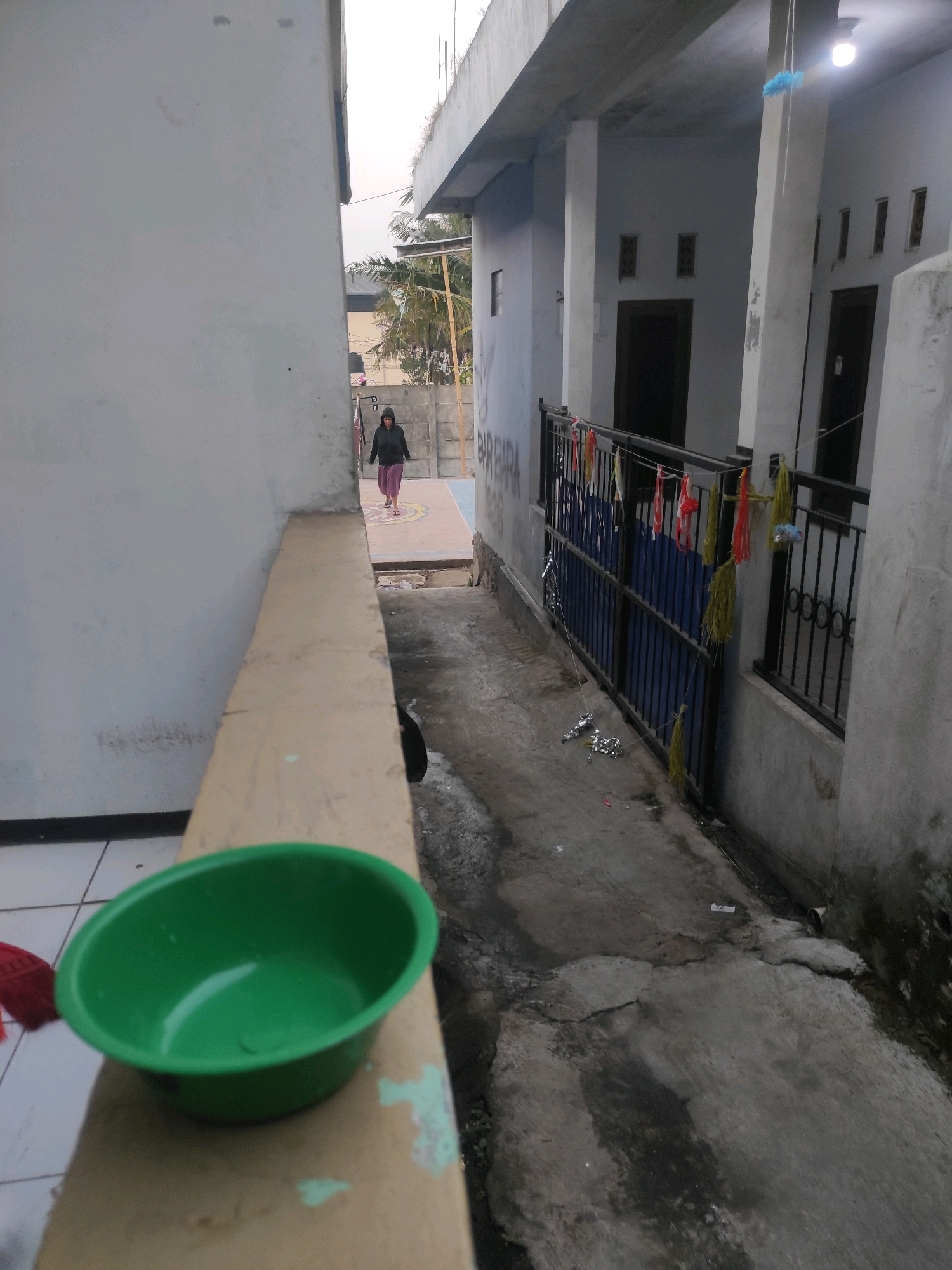 The image shows an outdoor scene in Padalarang, Indonesia, likely in a residential area. In the foreground, a green plastic basin sits on a beige ledge. To the left, a wall painted white. In the mid-ground, a person in a dark hooded jacket and purple pants walks away from the camera down a paved pathway. The pathway is narrow and flanked by buildings. To the right of the pathway, a black metal gate is visible, adorned with colorful tassels. Beyond the gate, the facade of a building with white walls and dark doors is seen. The sky appears overcast. The overall impression is a quiet, possibly residential, outdoor space.