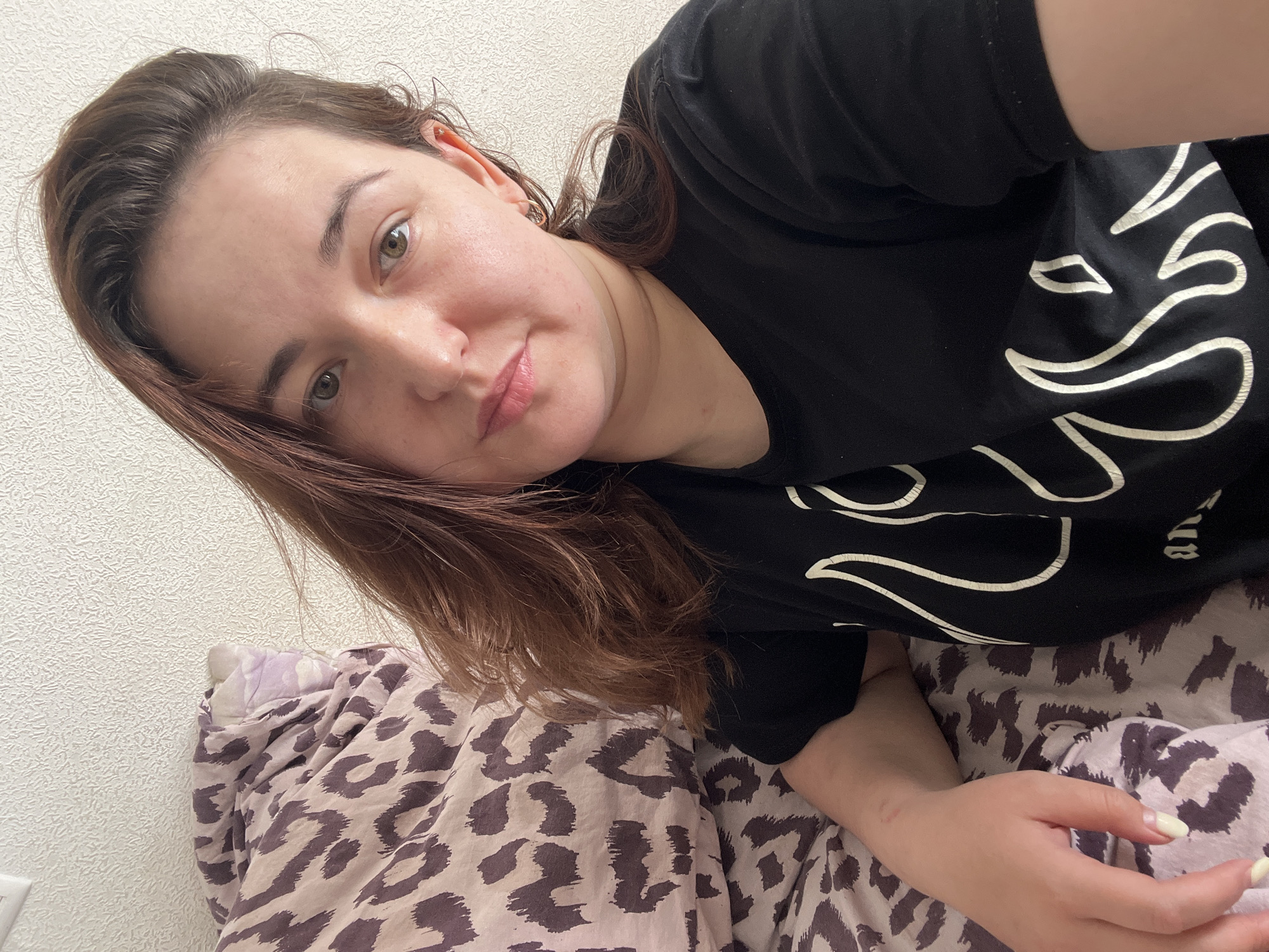 A young woman with medium-length brown hair and light brown eyes is lying in bed, looking at the camera. She is wearing a black t-shirt with a white flame-like design. A pillow with a leopard print pattern is visible behind her. The background appears to be a textured wall. This image was taken in Chelyabinsk, Russia.