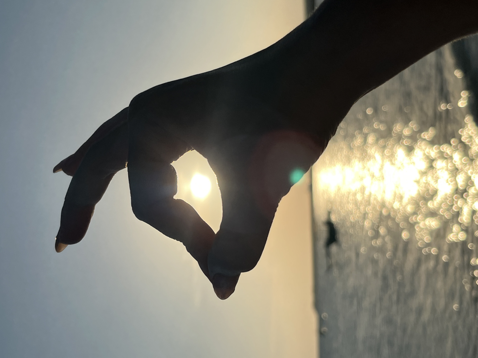 A close-up, silhouetted shot of a human hand forming an "OK" gesture against a sunset sky. The sun is visible through the center of the hand. The hand is in the foreground, with the ocean and a blurry horizon in the background. The water reflects the sunset, creating a shimmering effect of bokeh lights. A small, dark boat is visible in the distance on the water. The sky is a gradient of pale blue at the top transitioning to a warm, pale orange near the horizon. A subtle green lens flare is visible in the lower right portion of the image. The overall scene suggests a serene, natural environment, likely a beach in Kuta, Indonesia, during sunset.
