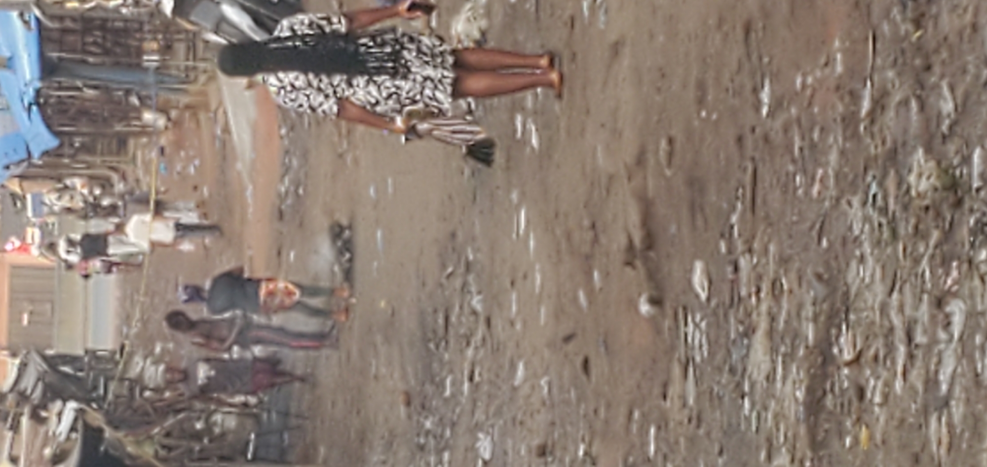 The image depicts a street scene in Onitsha, Nigeria. The ground is muddy and littered with debris. Several people are visible, including a woman in a patterned dress walking across the scene, and others standing or walking along the side of the road amidst vehicles and market stalls.