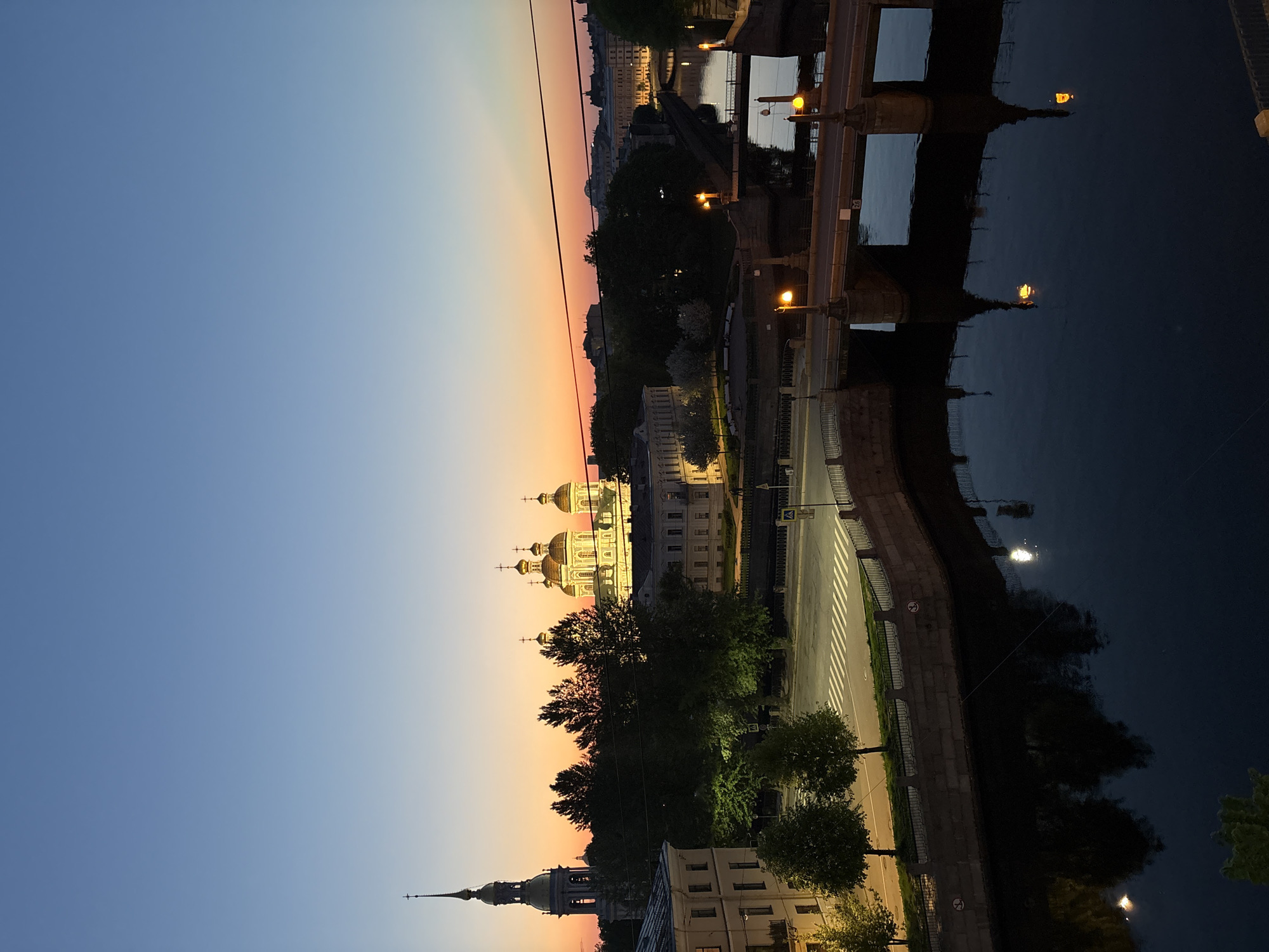 The image shows a cityscape at twilight, most likely taken in Saint Petersburg, Russia. In the foreground, a river reflects the lights from the city. A bridge, likely one of the many bridges in St. Petersburg, is visible with its lights illuminated. Along the riverbank, a street is lined with trees and buildings. Dominating the skyline is a golden-domed church, a common architectural feature in the city. A slender spire of another building is visible on the left side of the church. The sky transitions from a deep blue to a warm orange near the horizon. No people are visible in the image.