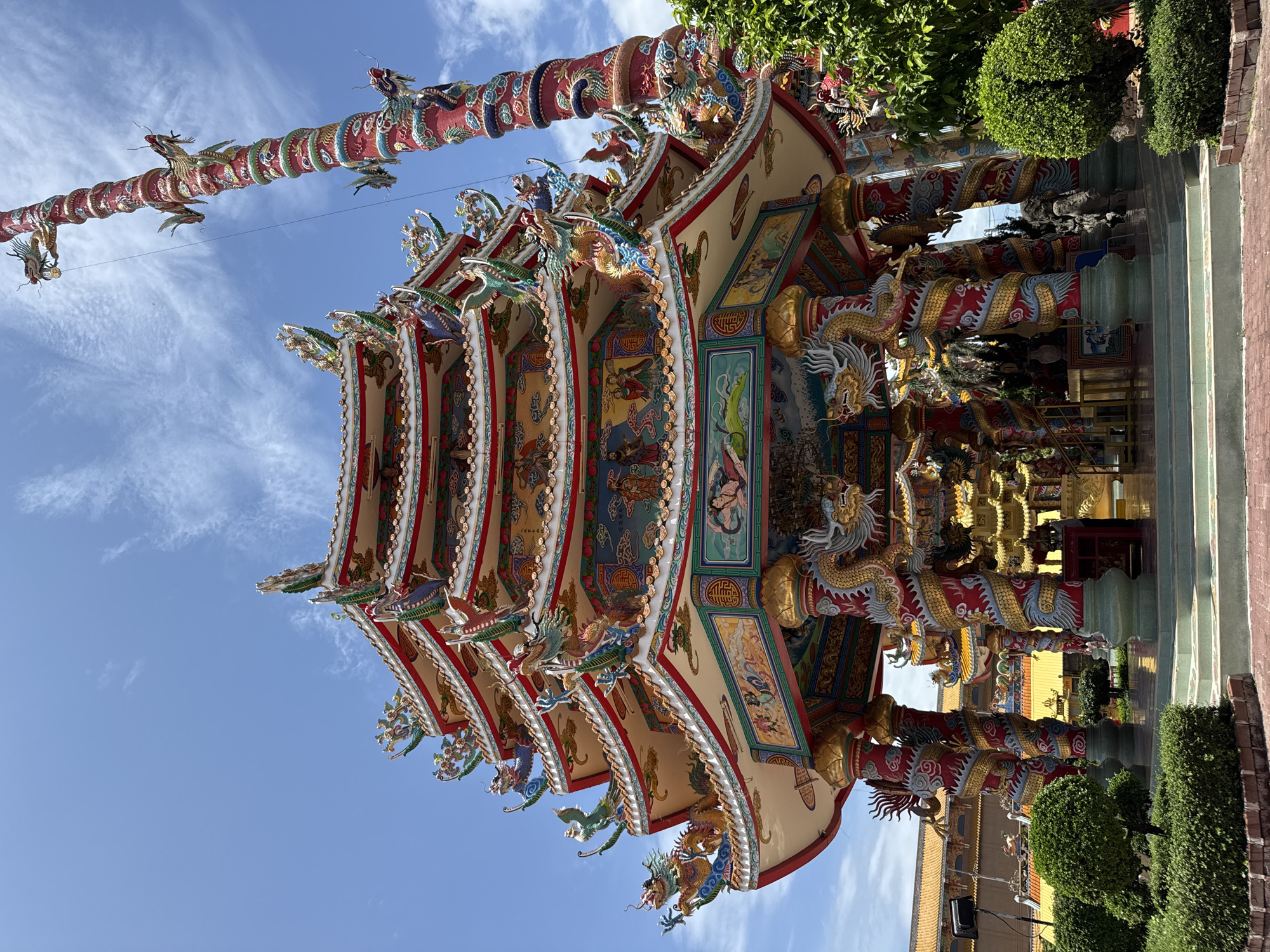 A multi-tiered, ornate temple structure with dragon motifs and brightly colored decorations stands under a clear blue sky with scattered clouds. A tall, red, dragon-entwined pillar is visible to the right of the temple. The temple is surrounded by lush green bushes and trees, with paved walkways and steps leading up to it. The background shows more temple structures with orange roofs. The scene is located in Ang Sila, Thailand. There are no people visible in the image.