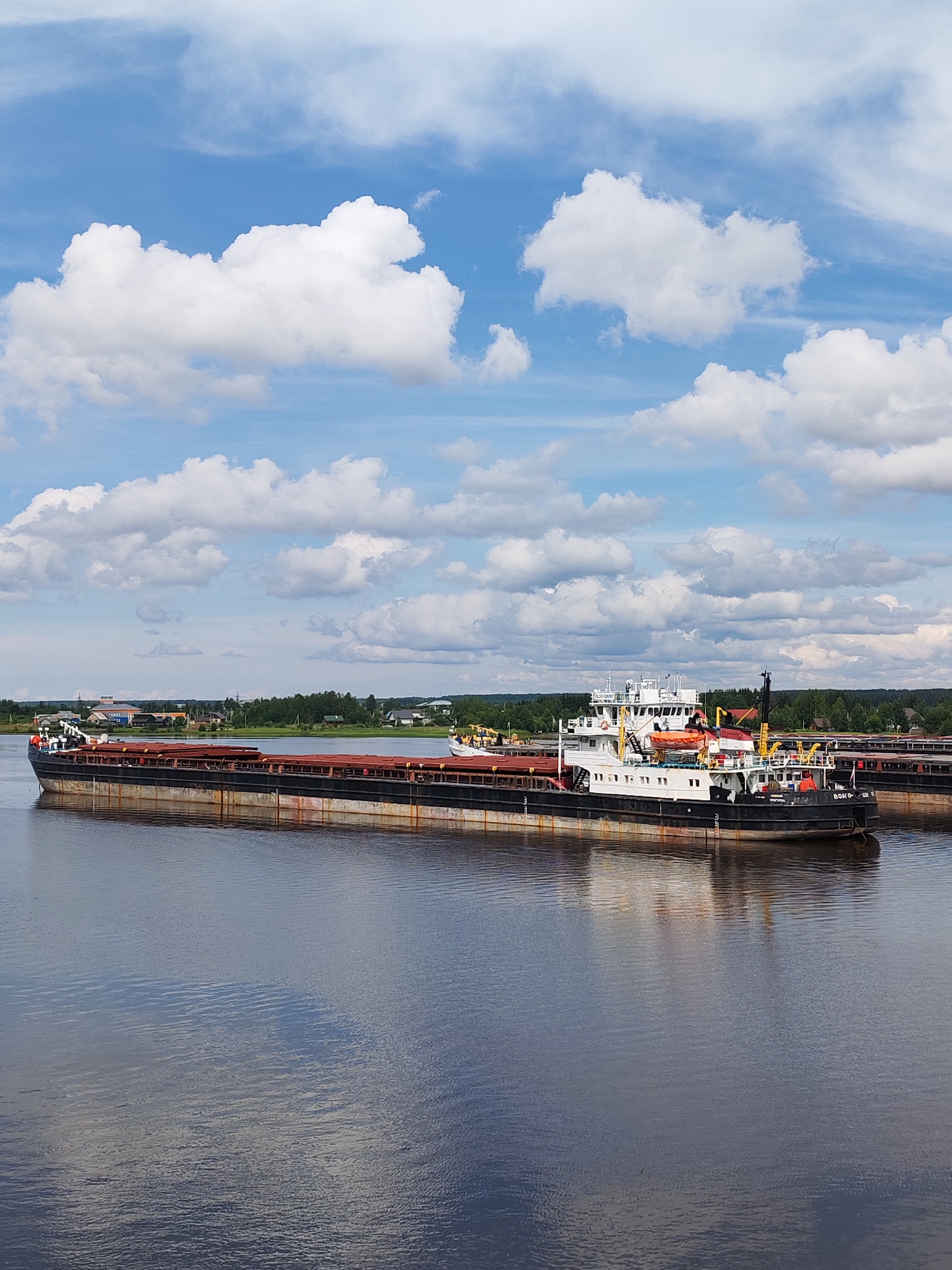 An aerial shot captures a wide river, with a large barge dominating the foreground. The barge is black and rust-colored, with a white cabin structure at its stern. It is loaded with what appears to be metal cargo. Another barge is partially visible behind it. The river is calm, reflecting the blue sky and white, fluffy clouds. The background shows a green treeline and a few buildings, suggesting a settlement along the riverbank. The image appears to be taken during the daytime. The location is identified as Shchetinino, Russia.