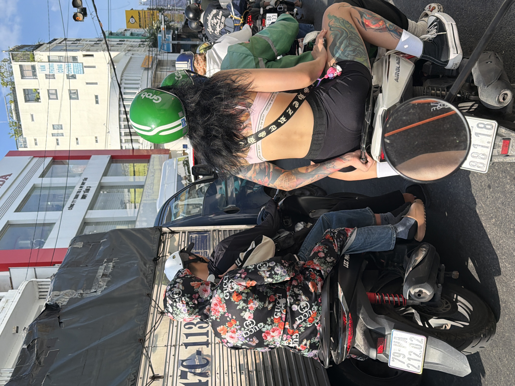 The image depicts a street scene in Nha Trang, Vietnam, characterized by dense traffic and urban architecture. In the foreground, two individuals are seated on a scooter. The person in the back, wearing a green helmet, has prominent tattoos on their arms and legs, and is dressed in a light pink sports bra and black shorts. The person in front wears a black hooded jacket with a floral pattern featuring Gucci branding, blue jeans, and light brown slides. They are both facing away from the camera, and the scooter they are on has a license plate that reads "79-XA 212.02". Behind them, a large truck with a tarp covering its cargo is visible, along with other vehicles and buildings that suggest a commercial or residential area. The overall impression is one of a bustling city street during daylight.