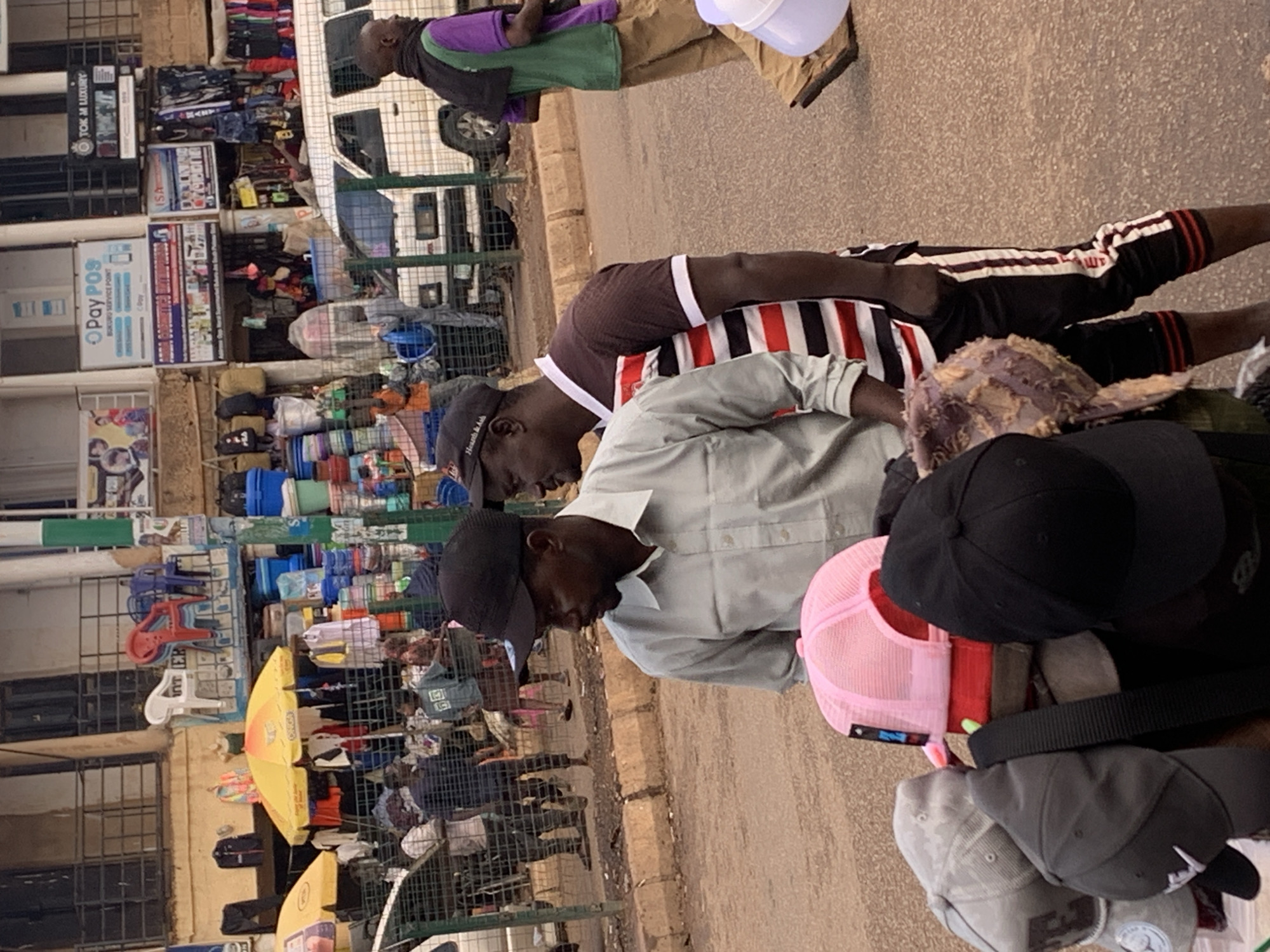 This image shows a street scene, likely in Bukuru, Nigeria. In the foreground, several people are visible, mostly men. One man is wearing a light-colored button-down shirt and a dark baseball cap. He appears to be looking down. To his left, another person is wearing a brown shirt with white and red stripes. Further back, a man in a purple and green vest stands near a white vehicle.
The scene appears to be a market or a street vendor area. Various objects are displayed, including stacks of colorful plastic containers, bags, and umbrellas. There are also some chairs, one red and one white, stacked on a metal grate. Some of the objects seem to be merchandise for sale. The ground is paved with a rough texture, possibly asphalt or concrete. The overall lighting suggests it could be daytime, with no strong indication of specific weather conditions.
Some visible text can be observed on signs in the background. One sign is for "Pay POS," another has "ISA" on it, and there are other signs with various lettering and logos, though most are not clearly legible. The atmosphere appears to be that of a busy commercial area.