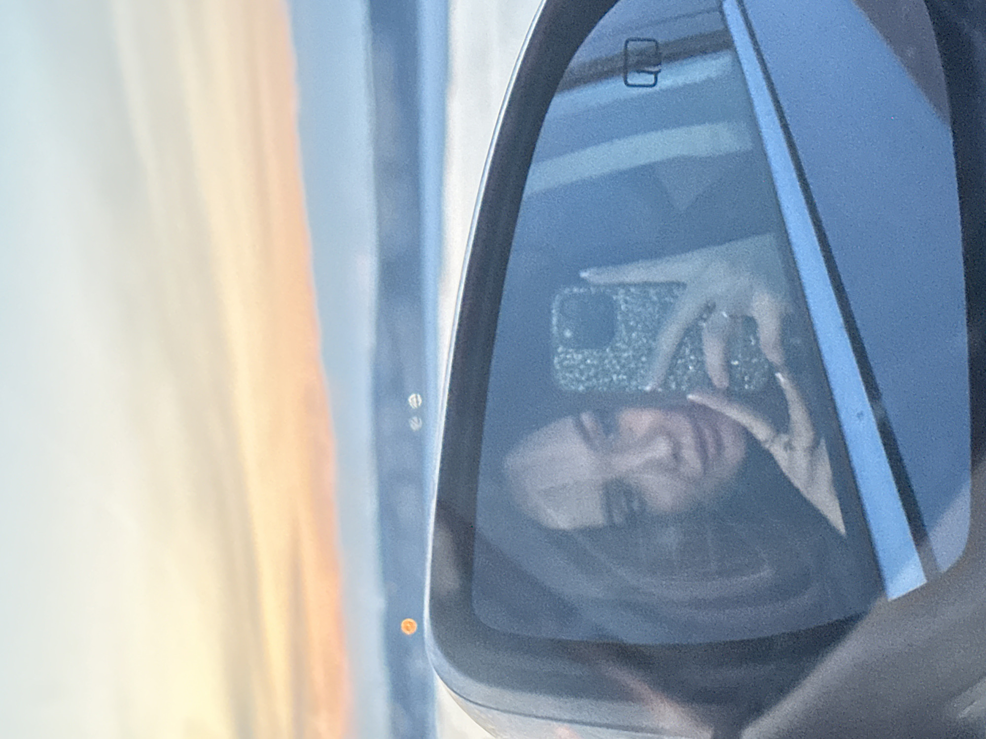 The image shows a person taking a selfie in a car's side mirror. The person has dark hair and is holding a phone with a glittery case. The mirror also reflects a blurred background of what appears to be the sky and possibly other cars. The location of the image is unknown.