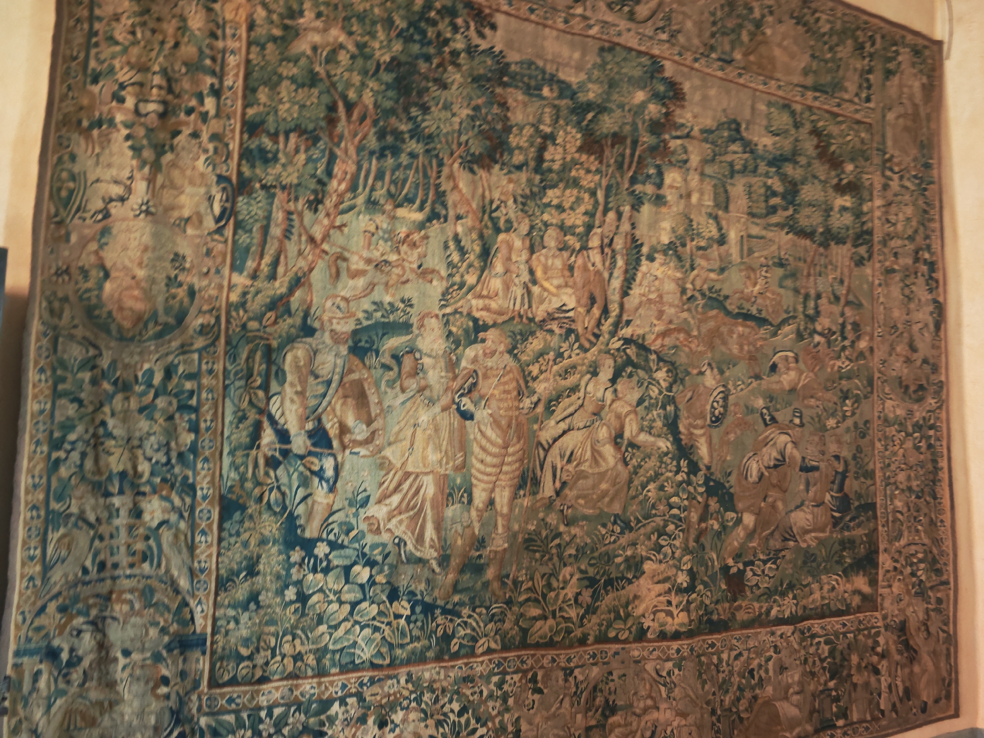 This is a photograph of a large, antique tapestry hanging on a wall, likely in an interior setting. The tapestry depicts a detailed scene with numerous figures in a forest or garden landscape. The colors are muted, with predominant tones of green, brown, and beige. Human figures are shown in period clothing, engaged in various activities, some appearing to be conversing or interacting in small groups. The landscape features trees with detailed foliage and a background that suggests depth. There is also a decorative border around the main scene. The tapestry is illuminated by ambient light, which casts subtle shadows and highlights.
The tapestry is located within Bauska Castle in Bauska, Latvia.