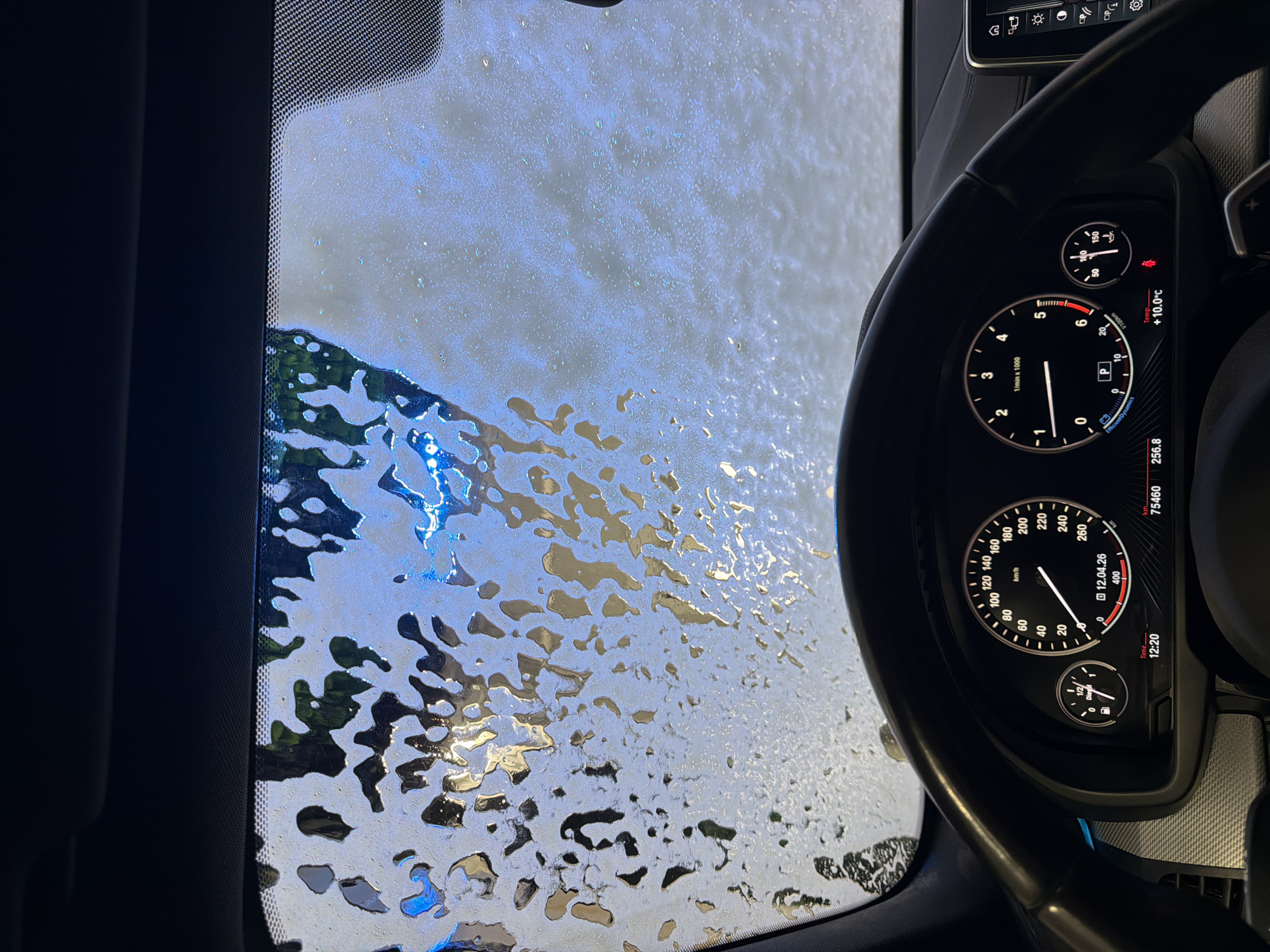 The image shows a view from inside a car going through a car wash. The windshield is covered in soap and water. The car's dashboard is visible, including the speedometer, tachometer, and other gauges. The speedometer reads 0 km/h, and the tachometer is near 0 RPM. The temperature is displayed as +10.0°C. The date on the dashboard is 12.04.26, and the time is 12:20. The odometer reads 75460 km, and fuel consumption shows 256.8. The car is in "P" (Park). The location is not indicated in the image.