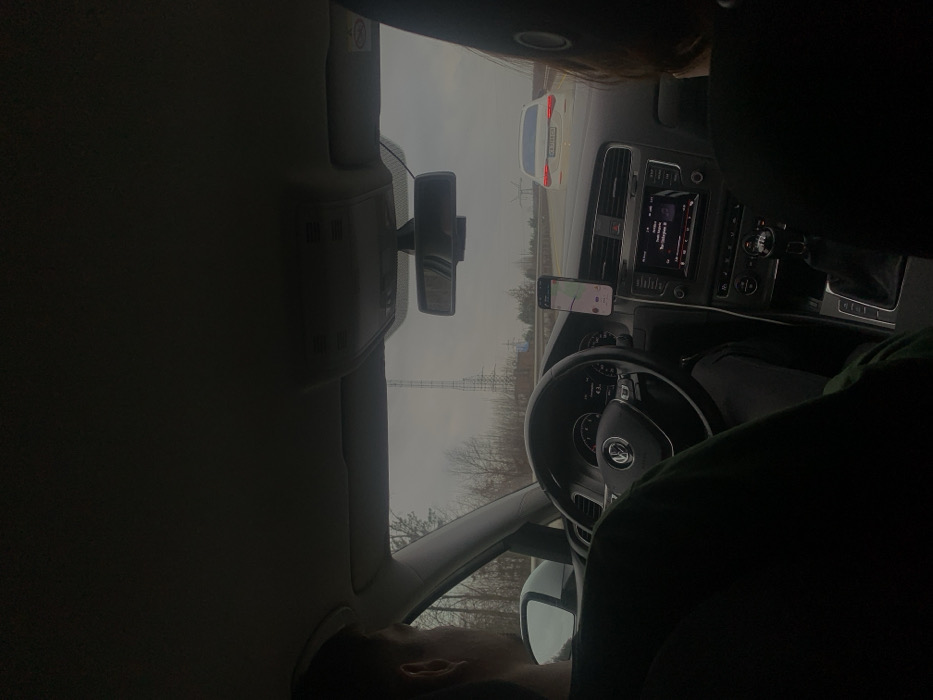 The image captures the interior perspective from the rear seat of a Volkswagen vehicle in Oliivka, Ukraine, looking forward through the windshield. A male driver, visible from the left side of his head and shoulder, wearing a dark top, grips the Volkswagen steering wheel. His left hand is positioned on the wheel. A front passenger, with dark hair, is partially visible from behind on the right side of the frame. A smartphone displaying a navigation application is mounted on the dashboard to the right of the steering wheel. The vehicle's infotainment system in the center console is active, showing a media interface. Beyond the windshield, a multi-lane road is visible under an overcast sky. A white sedan, with its brake lights illuminated, is directly ahead in the right lane. Bare-branched trees line the roadside, and a tall, lattice-style electrical or communication tower stands prominently in the distance to the left of the road. A smaller pylon-like structure is also visible further down the road on the right.
