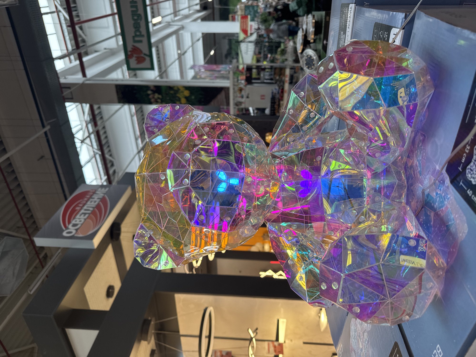 The image shows an indoor scene, likely within a large store. Two faceted, iridescent decorative items, possibly light fixtures or ornaments, are prominently displayed. These items are made of transparent material with a multifaceted, gem-like structure, reflecting a variety of colors, including blue, purple, and orange. The store environment is evident from the signage visible in the background. "Градини" and "ОСВЕТЛЕНИЕ" signs can be seen. The "ОСВЕТЛЕНИЕ" sign suggests the store may sell lighting products. Given the Cyrillic lettering on the signs, the location is likely in Bulgaria. Given the variety of items sold, it appears to be a large department store or home improvement center. There are no people visible in the immediate foreground, but blurred figures can be seen further in the background. Additional lighting fixtures and displays are partially visible in the background, reinforcing the store environment. Based on the signage visible, and the likelihood of the location being in Bulgaria, there is a high probability of the store being in Sofia, the capital city.