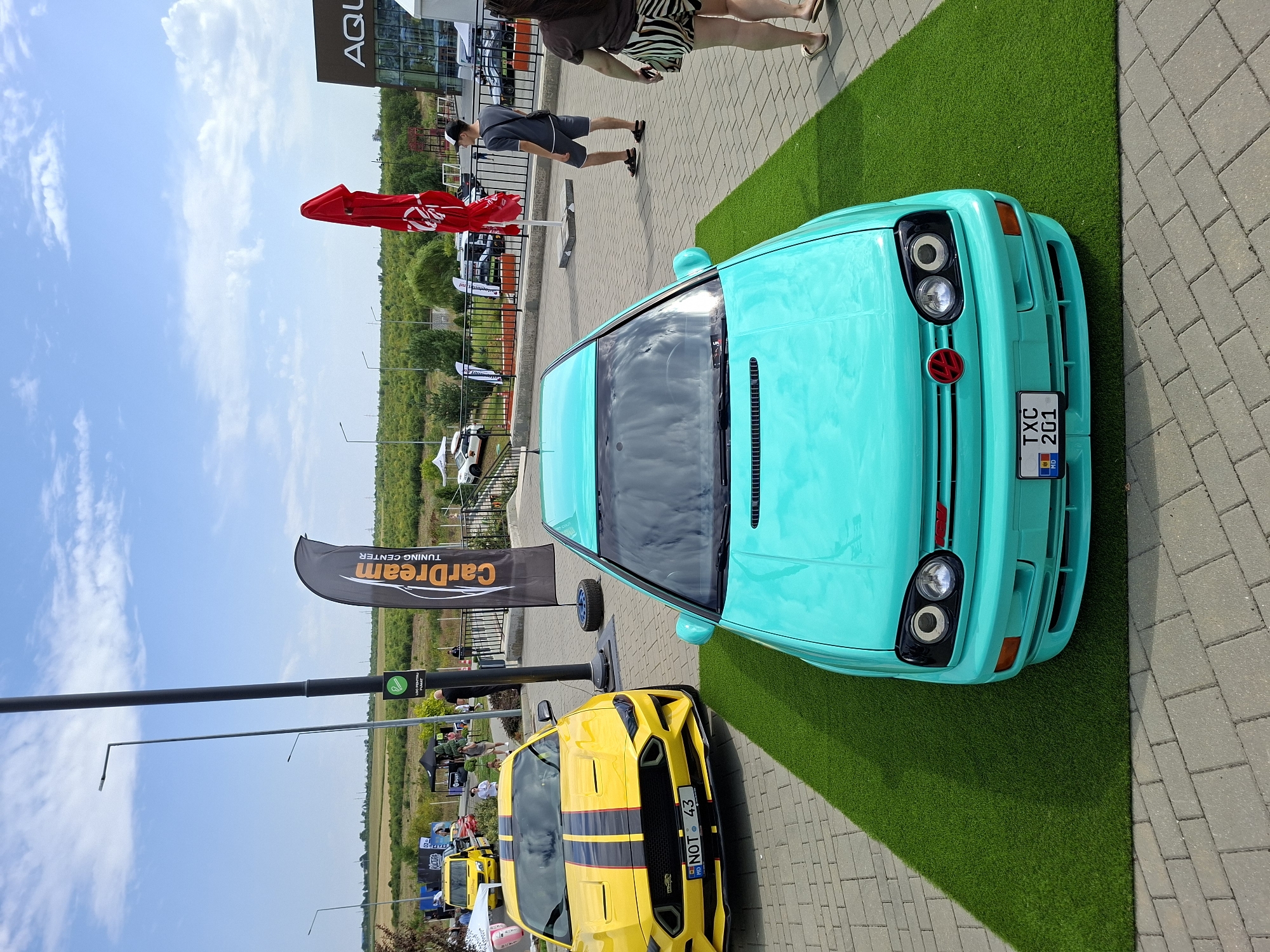 The image captures an outdoor event, likely a car show, under a partly cloudy sky. In the foreground, a turquoise Volkswagen Golf Mk3 is parked on a patch of artificial grass. The car features a custom red emblem on the grille and a license plate that reads "TXC 201" with the Moldovan flag. To the left of the Golf, a yellow Ford Mustang with black racing stripes is parked on a paved surface. A license plate on the Mustang reads "NOT 43 MD".
In the background, a man in a grey t-shirt and shorts stands near a black banner that says "CarDream TUNING CENTER". Further back, several tents and other vehicles are visible, including a white car with a roof rack. To the right, a building with the word "AQU" visible on its facade can be seen, and a bright red umbrella is unfurled. Several people are present, some walking and observing the cars. The overall setting appears to be a well-lit outdoor area with paved ground and some greenery in the distance, consistent with a location on the outskirts of Stăuceni, Moldova.