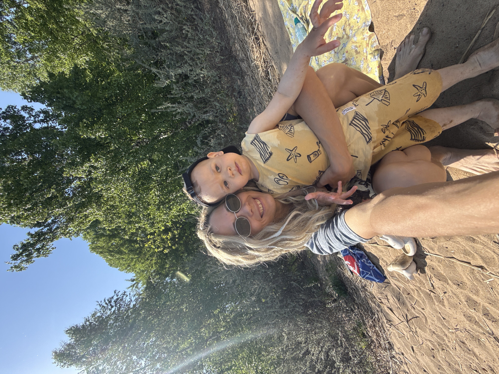 A woman wearing sunglasses and a blue and white striped shirt is lying on the ground with a child in her arms. The child is wearing a yellow outfit with black designs and a black baseball cap. They are both smiling at the camera. The background is a mix of sand, trees, and a blue sky. A blue and white item is next to the woman's arm. Given the location specified as Заволжье, Russia, this scene is likely taking place on or near a beach or sandy area near a body of water and trees in the region.
