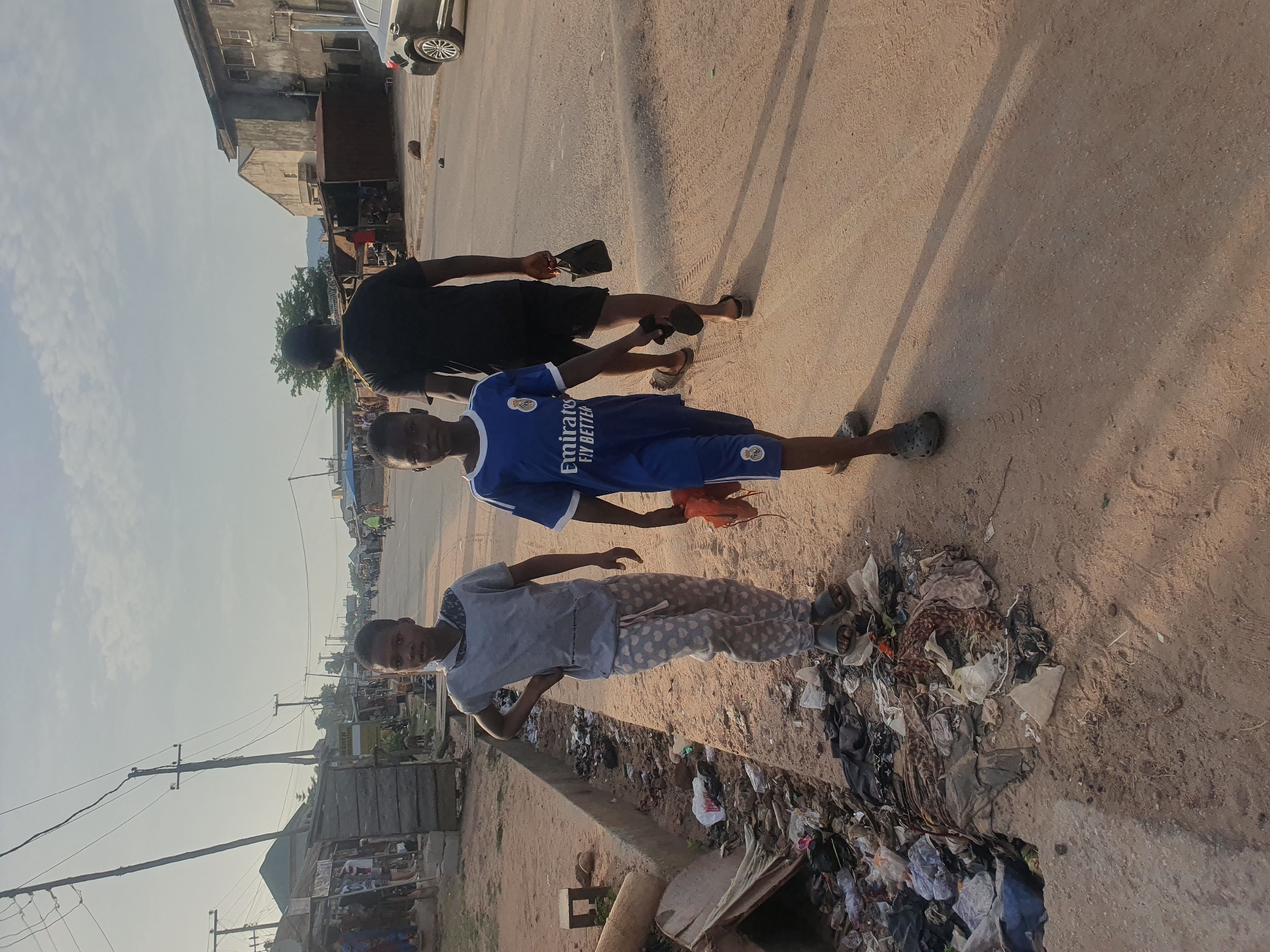 Three young males are walking on an unpaved road in Lokoja, Nigeria. A large pile of refuse and trash is located at the edge of the road on the right side. One male is wearing a blue soccer jersey and shorts. Another male is wearing a light-colored long-sleeved shirt and patterned pants. The third male is wearing a black shirt and shorts. A car is visible in the background, along with buildings and utility poles.