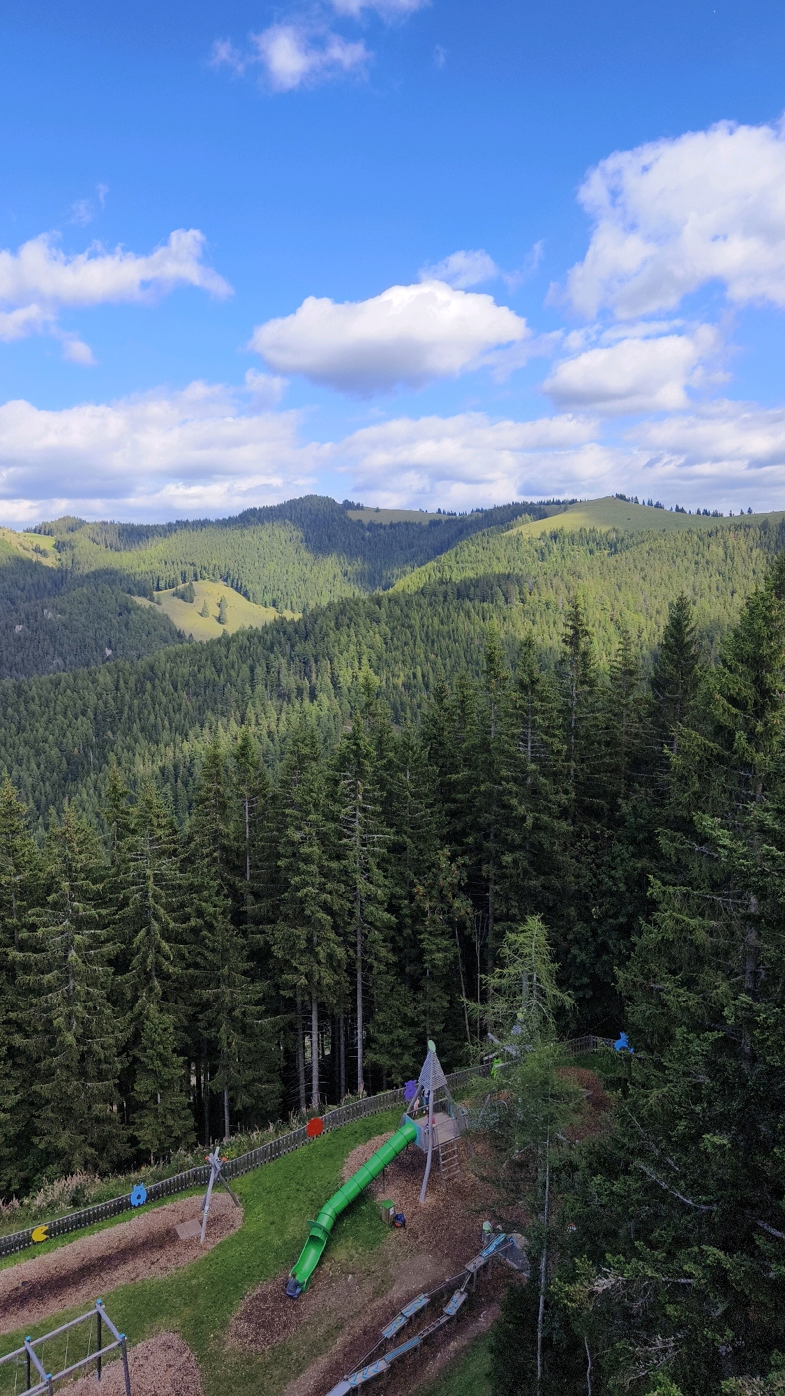 This is a high-angle, full shot of a playground nestled within a dense forest at the foot of rolling, tree-covered hills under a bright blue sky with scattered white clouds. The playground features a vibrant green tunnel slide, a wooden play structure with a roof, a swing set, and a zipline-like contraption with seats. The ground surfaces of the playground consist of wood chips and grass. The surrounding environment is dominated by tall, dark green evergreen trees, with patches of lighter green grassy clearings visible on the distant hills. Shadows cast by the clouds create a dappled light effect across the landscape. The image captures a serene, natural setting with a touch of playful activity.