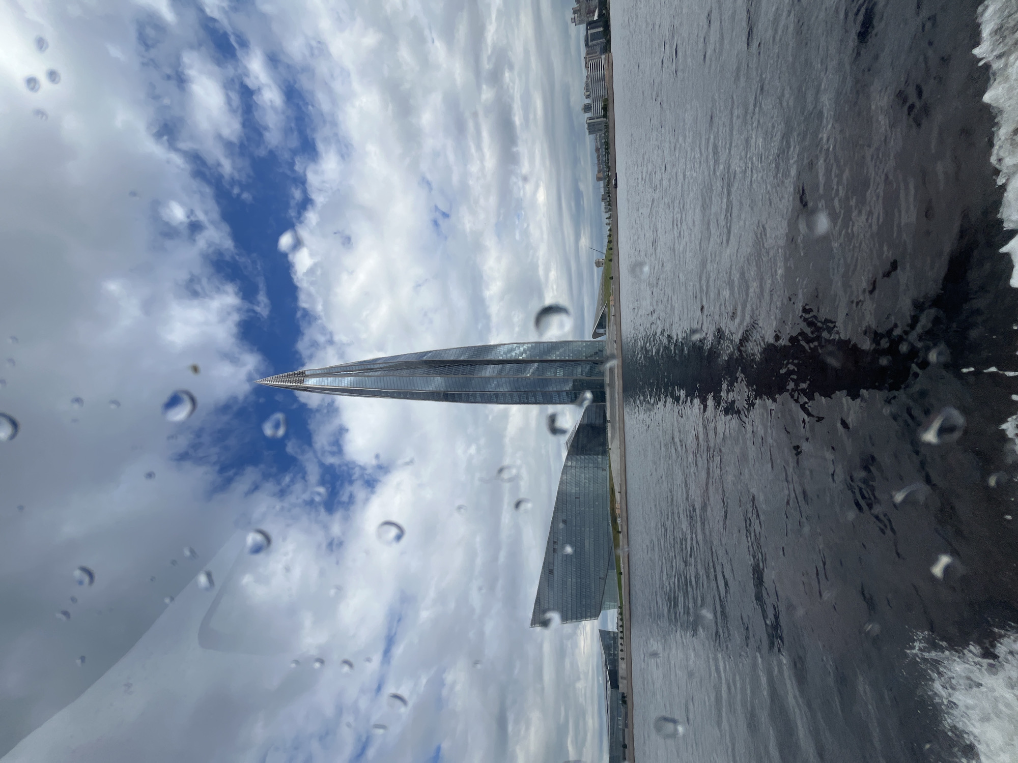 This image captures a view of the Lakhta Center in Saint Petersburg, Russia, from the perspective of a moving boat on the water. The Lakhta Center is a striking, contemporary skyscraper with a distinctive, twisting design, prominently featured in the center of the frame. To the left of the skyscraper is another modern, angular building with a glass facade, also part of the complex. The buildings are situated on the shore, with a body of water in the foreground. The water is dark and choppy, with a white wake visible at the bottom left, indicating the boat's movement. The sky above is a mixture of blue patches and white, dramatic clouds. Numerous water droplets are visible on the glass surface through which the photograph was taken, creating a slightly blurred and impressionistic effect. The overall scene suggests a cloudy, possibly humid day, with a clear view of the modern architectural marvels of Saint Petersburg.