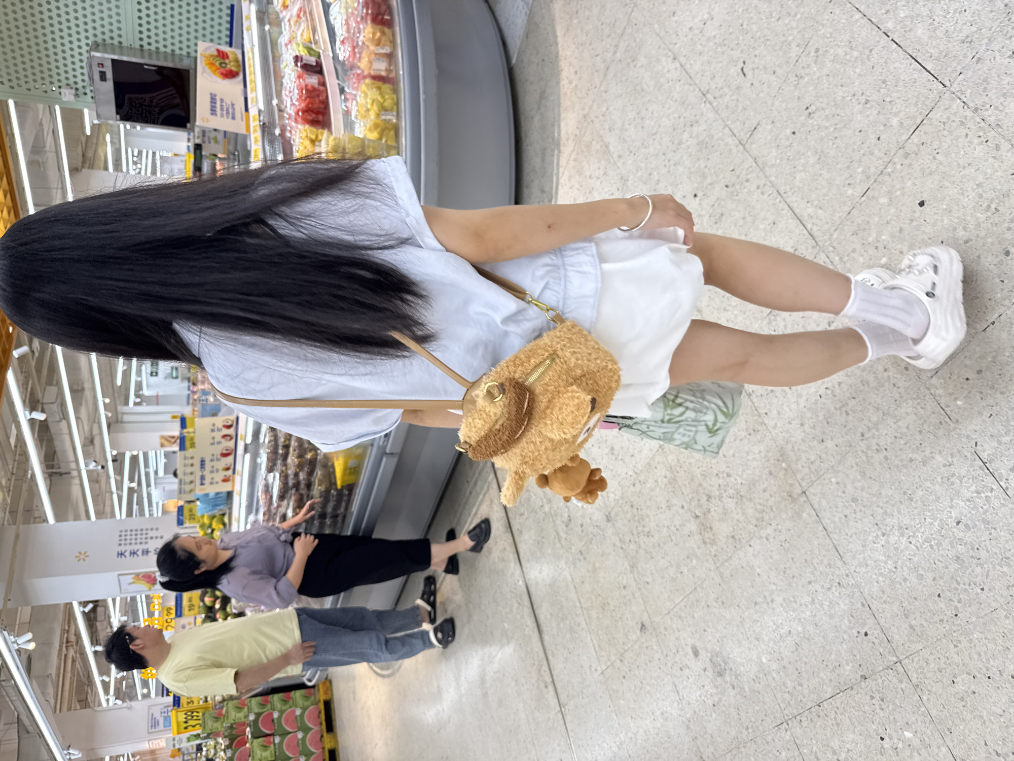 The image depicts a grocery store interior, likely in Huizhou, China, given the signage visible. In the foreground, a young girl with long black hair faces away from the camera. She is wearing a light blue short-sleeved shirt and a white skirt, complemented by white socks and white platform Crocs. A fuzzy, light brown bear-shaped crossbody bag hangs on her shoulder, with a small brown bear charm attached. She holds a white bag with green lettering in her left hand.
Further back, a woman in a light purple shirt and black pants stands near a refrigerated display case filled with fruits. To her left, a man in a yellow t-shirt and blue jeans stands looking towards the woman. The environment includes brightly lit aisles, colorful fruit displays in refrigerated cases, and tiled flooring. Signs with Chinese characters are visible throughout the store.