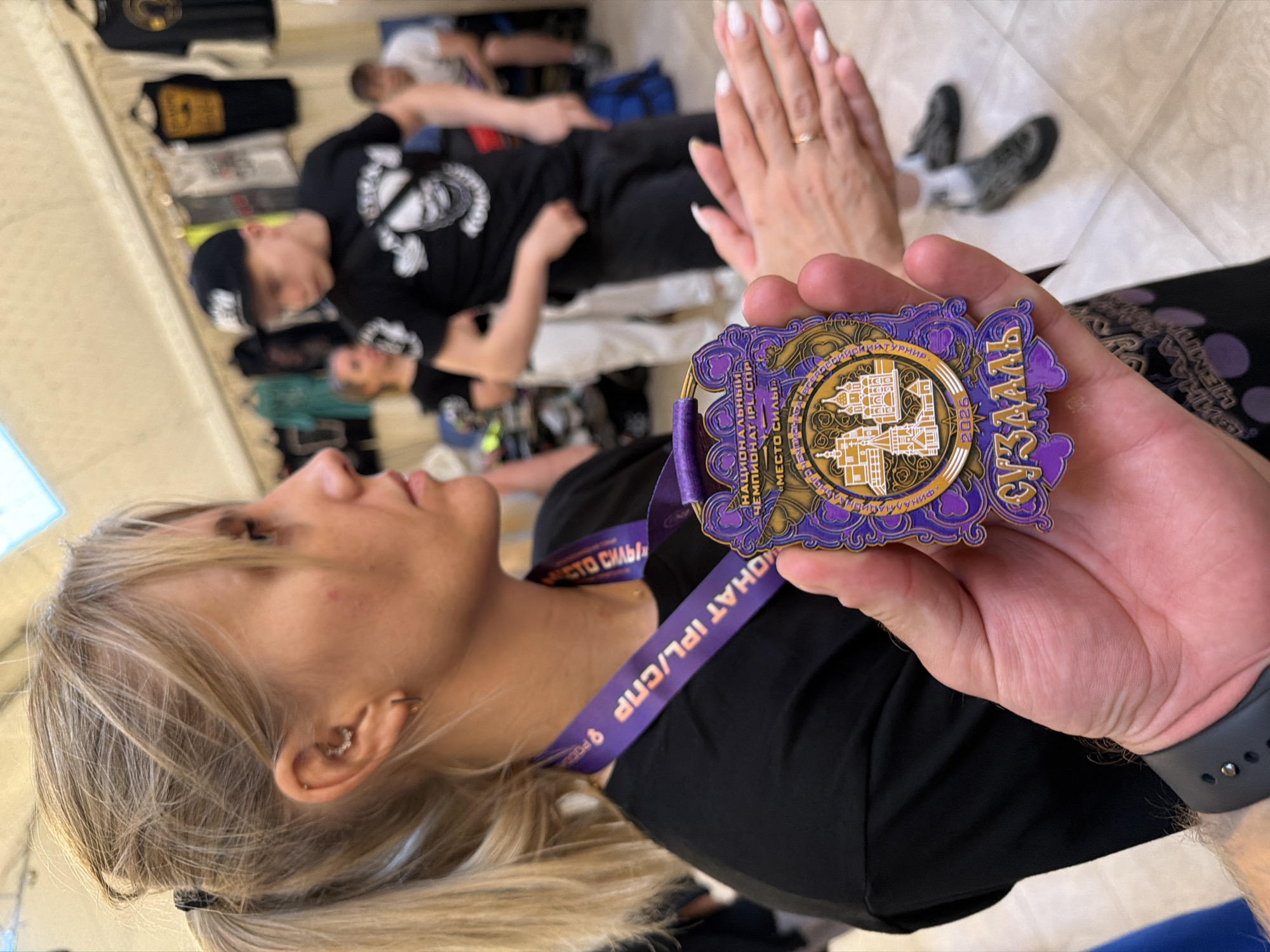 A close-up shot shows a woman's hand holding a medal. The medal is round with purple and gold embellishments and features images of churches. Text on the medal reads "НАЦИОНАЛЬНЫИ ЧЕМПИОНАТ IPL/СНР" and "СУЗДАЛЬ 2025". The woman is wearing a black t-shirt and a purple lanyard with similar text around her neck. In the background, a blurred group of people are visible, some wearing t-shirts and hats. The setting appears to be indoors, possibly a market or event, with racks of clothing visible in the distance. The floor is tiled. The woman's hand holding the medal has long, white-tipped fingernails and a gold ring on her finger.