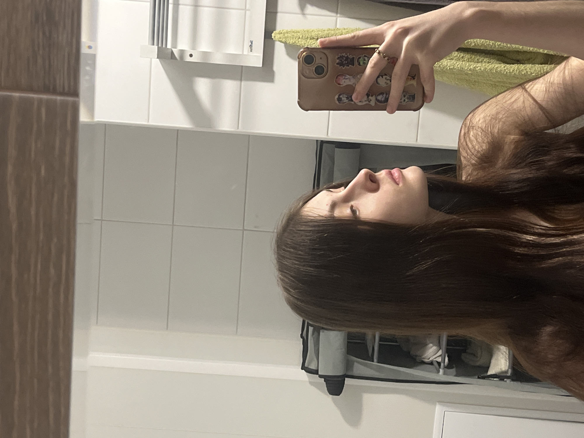 This vertical mirror selfie captures a person indoors, most likely in a bathroom. The setting features white tiled walls and artificial lighting.
The primary subject is a person with long, straight, dark brown hair, seen in profile. Only the right side of their face – nose, lips, and chin – is visible, along with their neck and shoulder. Their expression appears neutral, as they look slightly upwards while taking the photo.
The person holds a light brown or beige smartphone case, which is decorated with several small stickers or printed images depicting cartoon or anime-style characters. A ring with small, possibly beaded, details is worn on their right ring finger. Part of their arm is visible, showing fine arm hair.
Reflected in the mirror, behind the person, is a light green or yellowish textured towel draped over a metal rack with multiple horizontal bars. On the far left of the frame, a wooden element, possibly a door frame or cabinet edge, is visible. In the lower right corner, a gray cylindrical object and other background elements are subtly obscured by the person's hair. Faint, indistinct text, possibly "not," is reflected on the mirror in the upper left section. The overall impression is an everyday moment captured in an indoor setting. While the provided context suggests Saint Petersburg, Russia, there are no specific visual cues within the image to confirm this location.