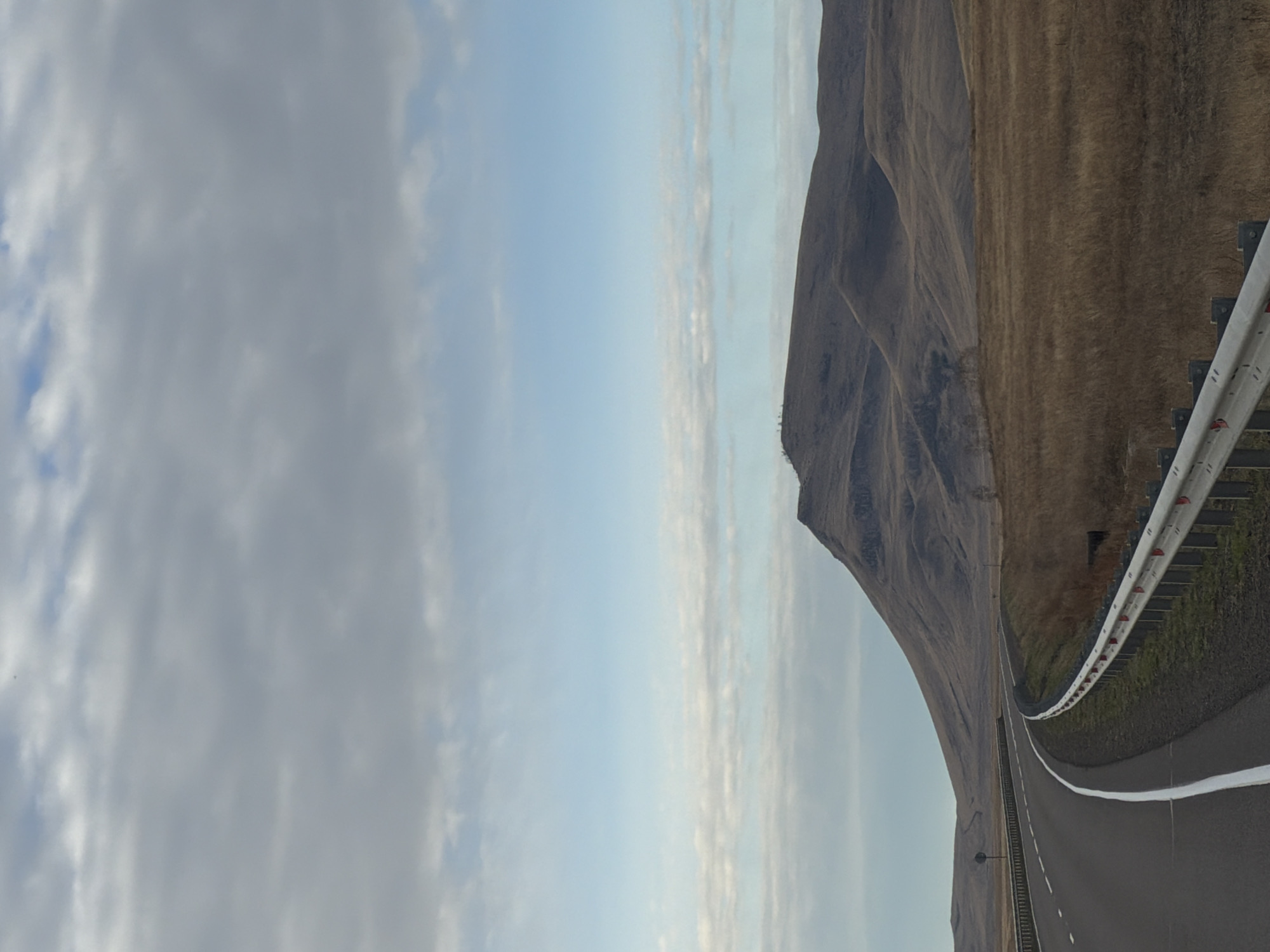 The image depicts a highway curving alongside a hillside. The road is asphalt with white lane markings and a guardrail. The hillside is covered in dry vegetation. The sky is partially cloudy. The image is rotated 90 degrees counter-clockwise. The location is Сарагаш, Russia.
