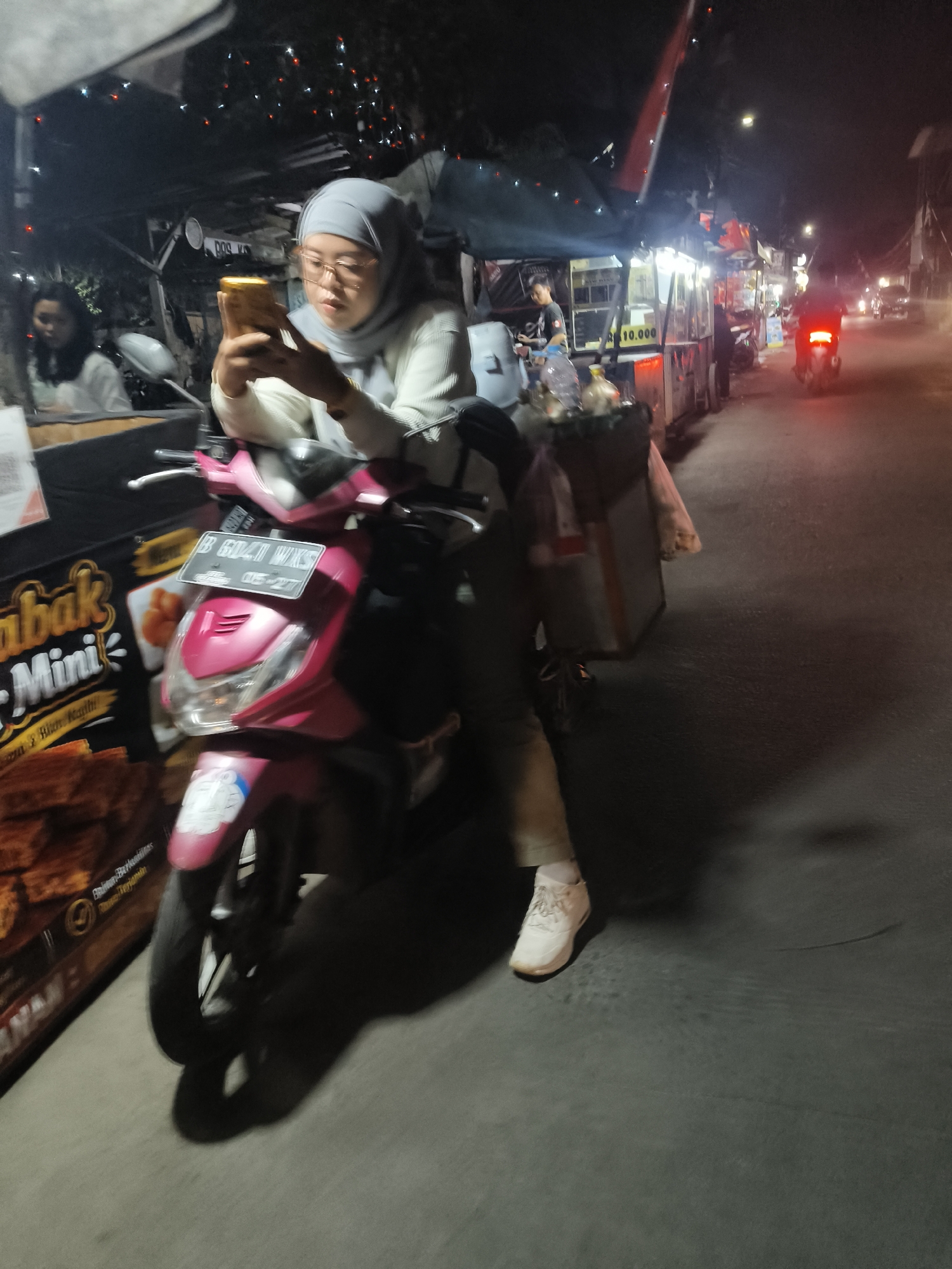 A medium shot captures a young woman wearing a grey hijab and glasses, sitting on a pink scooter at night on a street in South Tangerang, Indonesia. She is holding a gold-colored smartphone and appears to be looking at it. The scooter has a license plate that reads "B 6047 WXS". Behind her, there are food stalls with lights and a person working at one of them. A red motorcycle is visible in the distance. The woman is wearing a light-colored long-sleeved shirt and grey pants, with white sneakers. A black box is attached to the back of the scooter. The environment suggests a bustling street scene with food vendors and ongoing traffic.