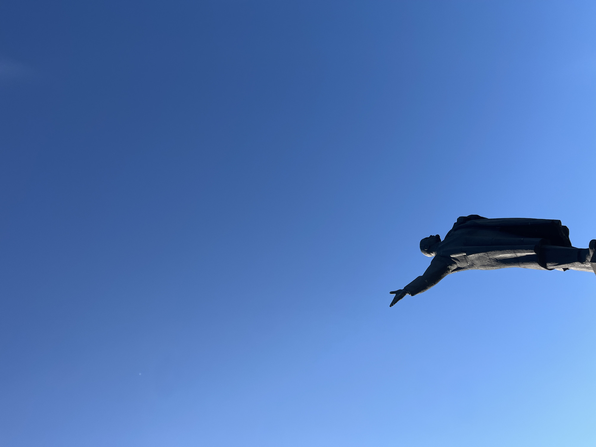 The image shows a statue of a man in a suit and coat, with his right arm raised and pointing upwards. The statue is dark and appears to be made of metal. It is silhouetted against a clear blue sky. The statue is positioned at the bottom right of the frame, with the majority of the image being the sky. There is a tiny, faint white speck visible in the sky to the upper left of the statue, which might be a distant aircraft or a cloud. The statue is likely located in Nizhny Novgorod, Russia, given the context provided in the prompt.