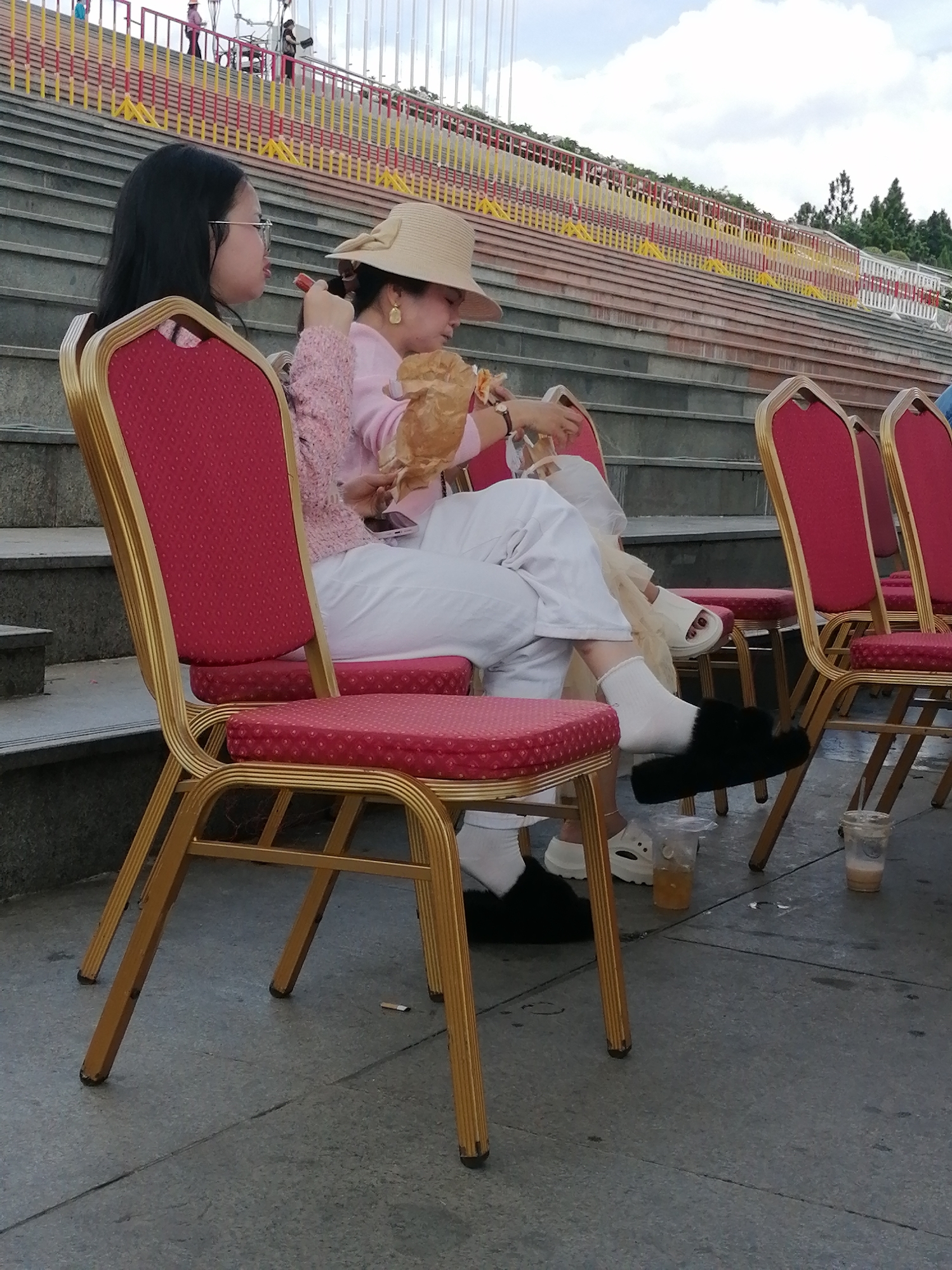 Two Vietnamese women are seated on red upholstered chairs with gold metal frames, facing a stepped seating area. The woman on the left wears glasses, a pink knitted sweater, and white pants. She holds a small object, possibly food, in her hand. The woman on the right wears a wide-brimmed hat, a pink sweater, and white pants. She is holding a crumpled paper bag and appears to be eating or holding something from it. Both women are wearing white socks. The woman on the left is wearing black furry slippers, while the woman on the right is wearing white slide sandals. They are outdoors, with a stone-like ground surface. In the background, there are tiered concrete steps with red and yellow railings, suggesting an amphitheater or outdoor seating area. The sky is partially cloudy. The scene appears to be at the Da Lat Flower Garden in Da Lat, Vietnam, a popular tourist attraction known for its floral displays and outdoor event spaces.