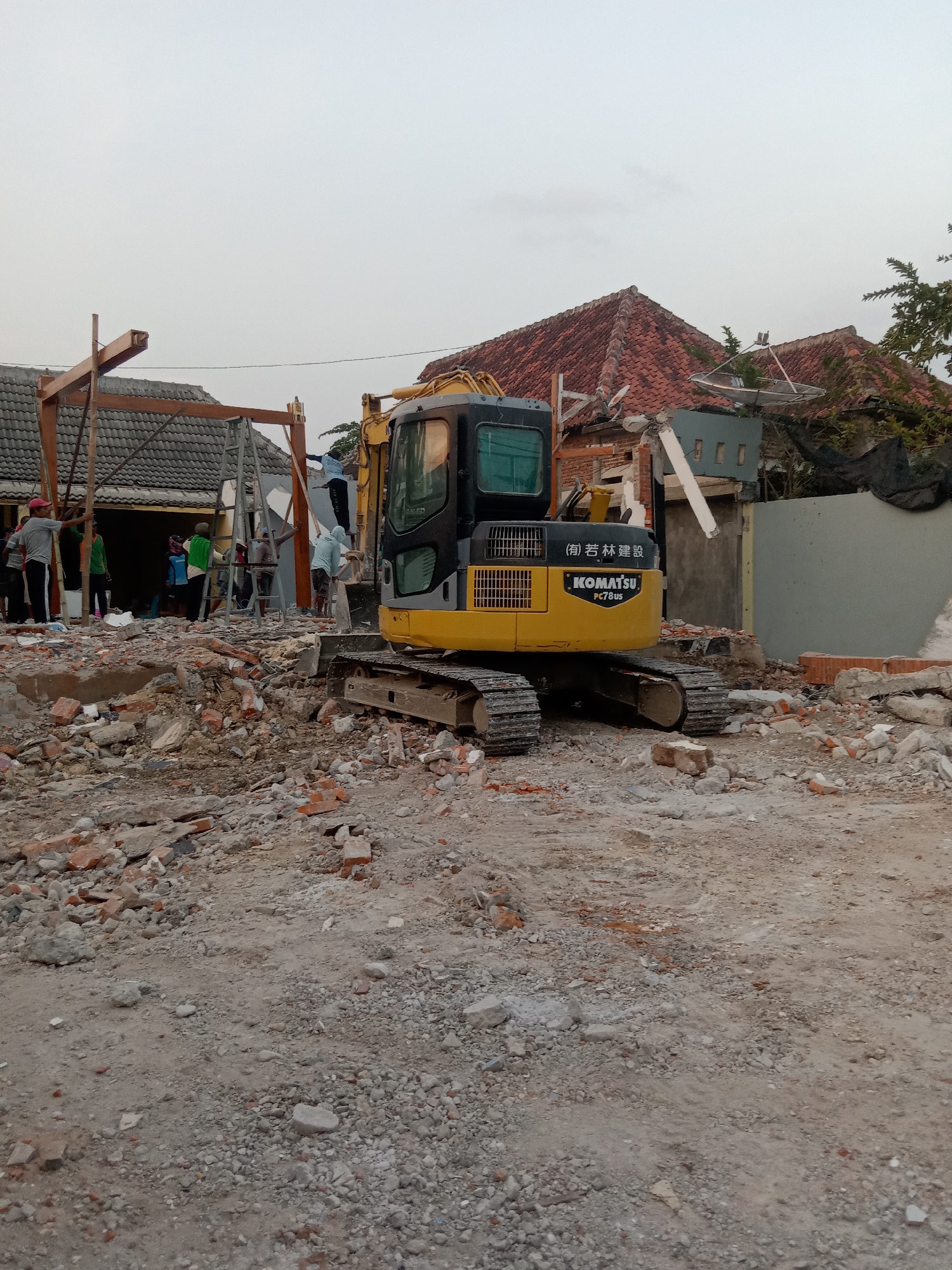 In Stiaji, Indonesia, a construction site is depicted with an excavator in the foreground. The excavator is yellow and black, with "KOMATSU PC78US" visible on its side. It is positioned amidst a pile of rubble, which consists of broken bricks, concrete, and other debris. In the background, a partially demolished building with a red tiled roof is visible. Several people, dressed in casual clothing, are scattered around the site, some appearing to be working on the demolition or cleanup. The environment is dusty and appears to be an outdoor setting under an overcast sky.