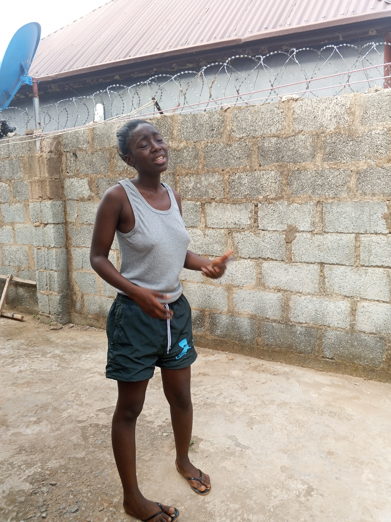A woman stands in front of a cinderblock wall topped with razor wire. She is wearing a gray tank top and dark green shorts. Her expression appears emotional. A satellite dish is visible on the left, attached to a building with a brown roof. A wooden ladder leans against the wall on the left. The ground appears to be concrete or packed dirt. The location is potentially within a compound or secured area in Abuja, Nigeria, indicated by the security measures of the wall and razor wire.