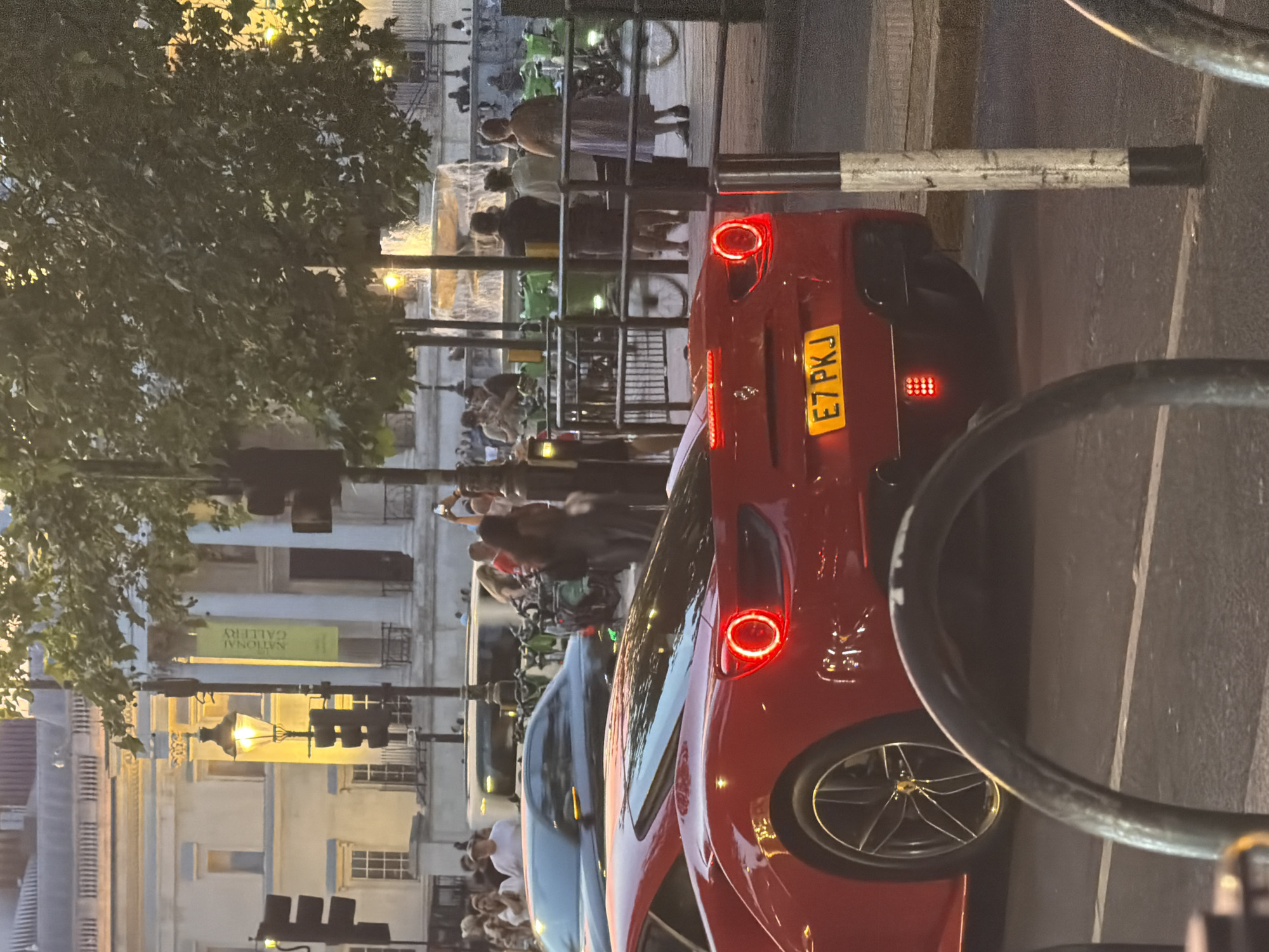 This image depicts a street scene in London, United Kingdom. In the background is the National Gallery. A crowd of people is gathered near a railing, with bicycles visible. In the foreground, a red sports car is parked on the side of the road. The car's license plate reads "E7 PKJ". A bicycle rack is partially visible, positioned close to the car. Traffic signals and street lamps are present, suggesting it is dusk or night. The overall environment suggests a tourist area or public space within the city.