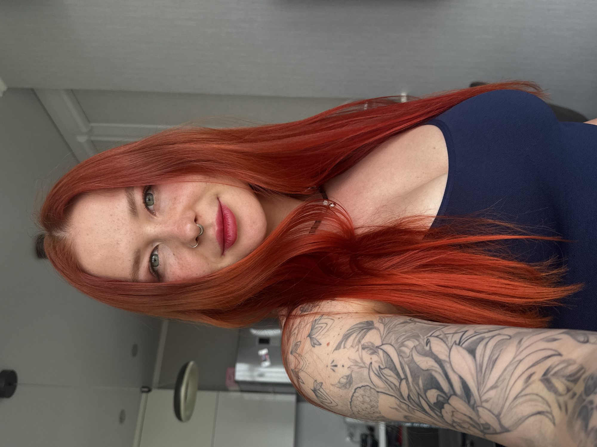 A woman with red hair, fair skin, and freckles poses for a selfie in what appears to be an indoor environment. She is wearing a dark blue top. She has a nose piercing and red lipstick. Her right arm is visible, covered with a detailed black and grey tattoo. The background suggests a domestic setting, possibly a kitchen, with white cabinets visible. The image appears to have been taken in a bright, well-lit space. There is no indication that this photograph was taken in Rostov-on-Don, Russia.