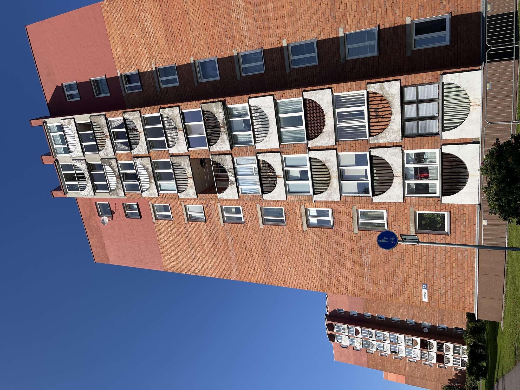 A low-angle, exterior shot captures two imposing, multi-story brick apartment buildings under a clear blue sky in Kohtla-Järve linn, Estonia. The building in the foreground, constructed from reddish-brown brick, features numerous windows and a series of arched, concrete balconies with dark metal railings. A portion of the upper facade of this building is painted a contrasting salmon pink. A street sign reading "Kalevi 22" is visible on the brick wall. To the left, a blue circular sign with a pedestrian and bicycle icon is mounted on a pole. The second apartment building, partially visible behind the first, shares similar architectural elements, including the brick construction and balconies. The scene is devoid of any visible people, pets, or vehicles. The foreground includes a grassy area and a sidewalk, with some low green bushes and shrubbery. The overall impression is a residential urban environment.