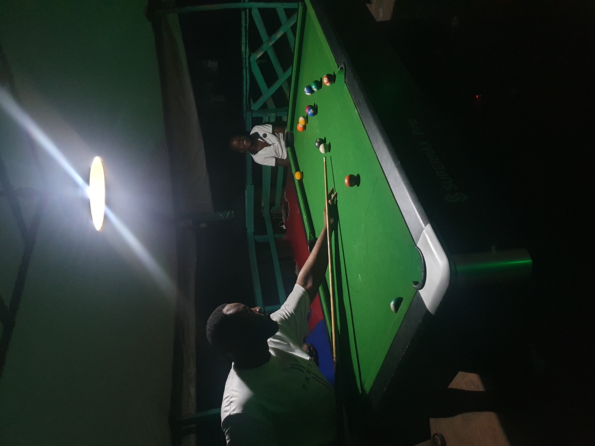 Two individuals are engaged in a game of pool at an outdoor, covered establishment, likely in Lokoja, Nigeria. In the foreground, a man wearing a white t-shirt is in the process of striking a pool ball with a cue stick. The pool table is green with a standard arrangement of colored balls. In the background, another individual, a boy or young man, is seated and smiling, watching the game. The environment appears to be dimly lit, with a single overhead light illuminating the pool table. The structure suggests an open-air recreational area, possibly a bar or lounge. The overall scene depicts a casual and social moment of leisure.