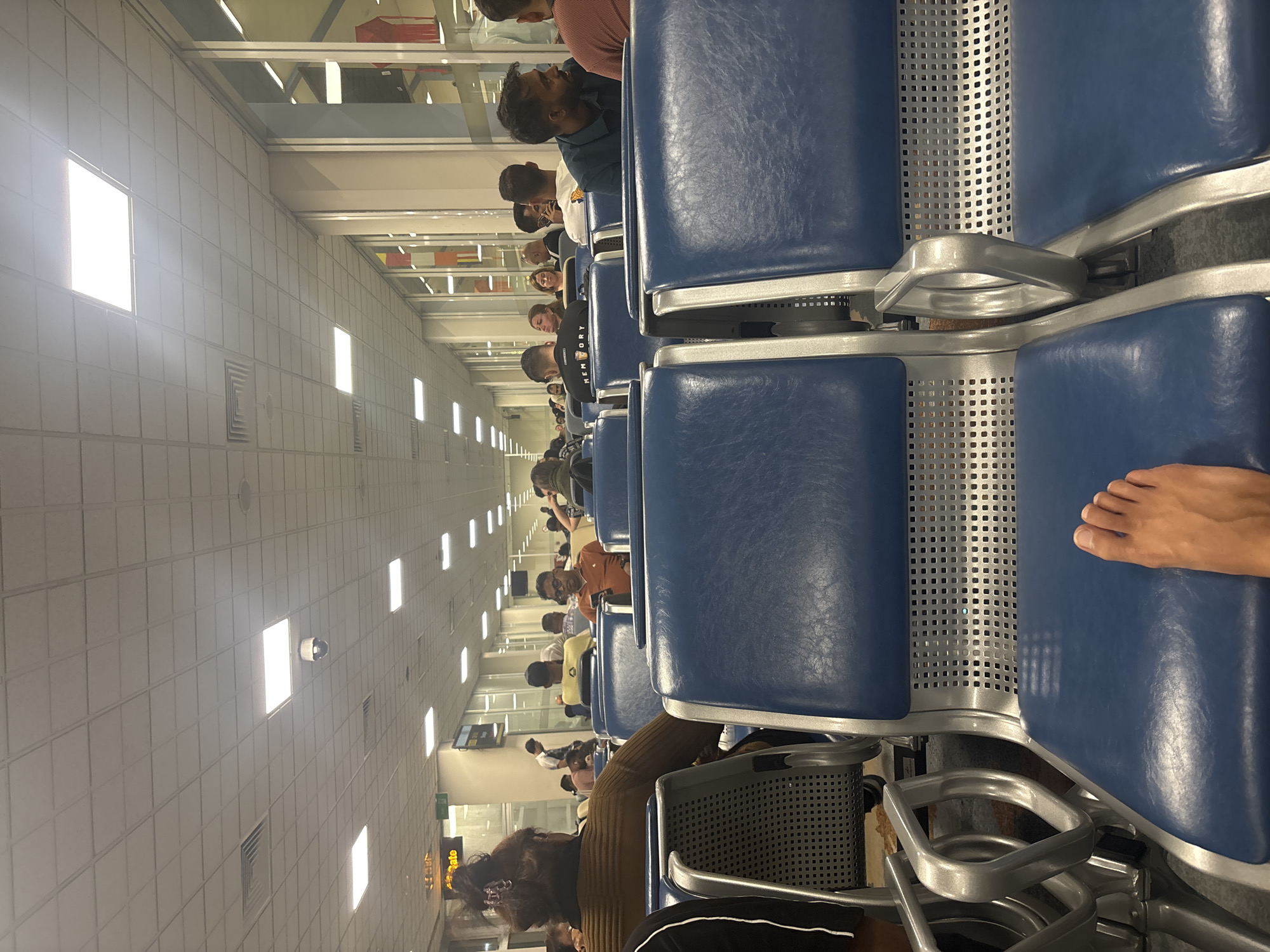 This image captures an indoor public waiting area in Katunayake, Sri Lanka, consistent with an airport terminal setting. The perspective is from a low vantage point, showcasing the bare right foot of the photographer resting on a blue upholstered seat in the immediate foreground.
The scene features multiple rows of standardized public seating units, characterized by blue padded seats and backs, silver-colored metallic frames, and perforated metal backrests. Numerous individuals are seated throughout these rows, with additional people standing further back. In the midground, a person with dark hair styled in a bun, wearing a brown long-sleeved top, is seen from behind on the left. Adjacent to them, another individual wears a black t-shirt with "MEMORY" printed in white. To the right, a man with dark skin and eyeglasses, dressed in an orange polo shirt, is focused on a smartphone in his hands. Further right, a bearded man in a dark blue polo shirt is seated, looking towards the right side of the frame. A staff member in a light blue uniform shirt stands near a pillar in the background.
The ceiling is a grid of white square acoustic tiles, illuminated by numerous recessed square LED panel lights. A white dome-shaped surveillance camera is centrally mounted on the ceiling. Ventilation grilles are also visible. On the far left, a yellow sign displaying "Gate" and other partially obscured text is present. The right side of the frame features large vertical glass panels, reflecting interior elements and revealing indistinct colorful objects beyond. The floor appears to be a light-colored tiled surface. The primary activity observed is people waiting and utilizing personal electronic devices.