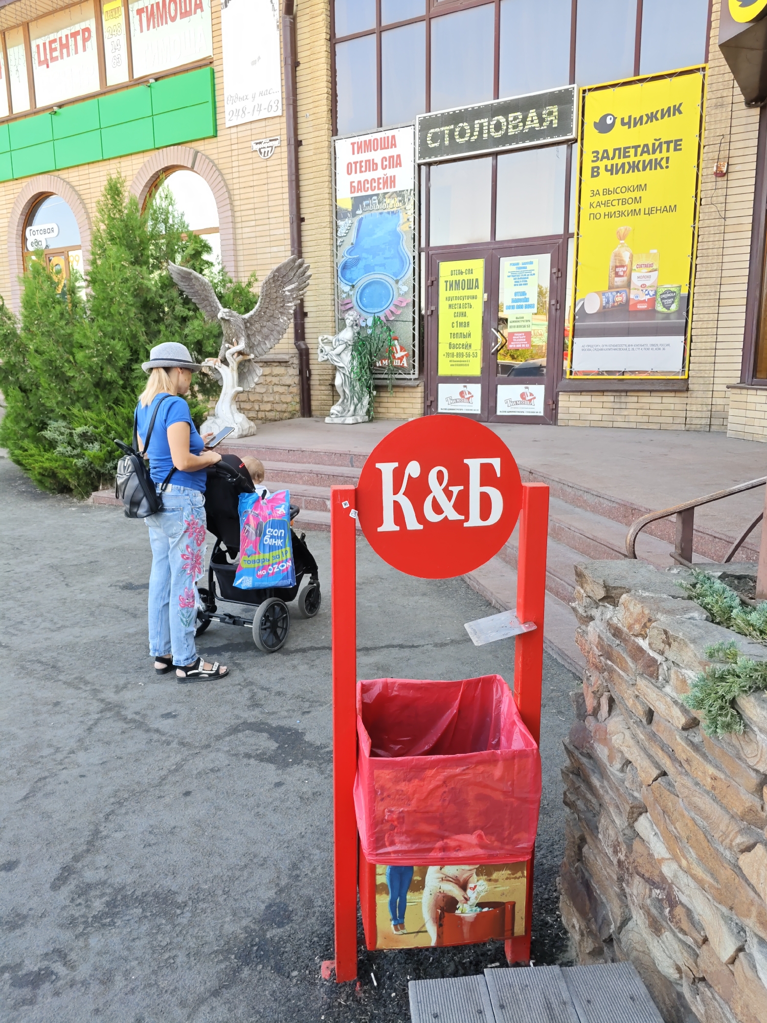 The image shows a street scene in front of a commercial building. In the foreground, a red trash receptacle with a white "K&Б" logo is visible. To the left of the receptacle, a woman in a blue shirt and floral-patterned jeans is standing next to a baby stroller. She is wearing a hat and sandals, and she appears to be looking at her phone. A blue shopping bag with "ozon" printed on it is in the stroller. The building behind them has large windows and signs, including one that reads "СТОЛОВАЯ" (Dining room) in large illuminated letters, and another yellow sign with a bird logo and Russian text that translates to "Chizhik, fly in to Chizhik! For high quality at low prices." The building appears to be a commercial establishment. The overall setting suggests an outdoor area in a city, potentially with some retail or service businesses. Based on the presence of signs in Russian and the architectural style, the location is likely in Russia. The image does not contain any specific visual cues that definitively place it in Rostov-on-Don, Russia.