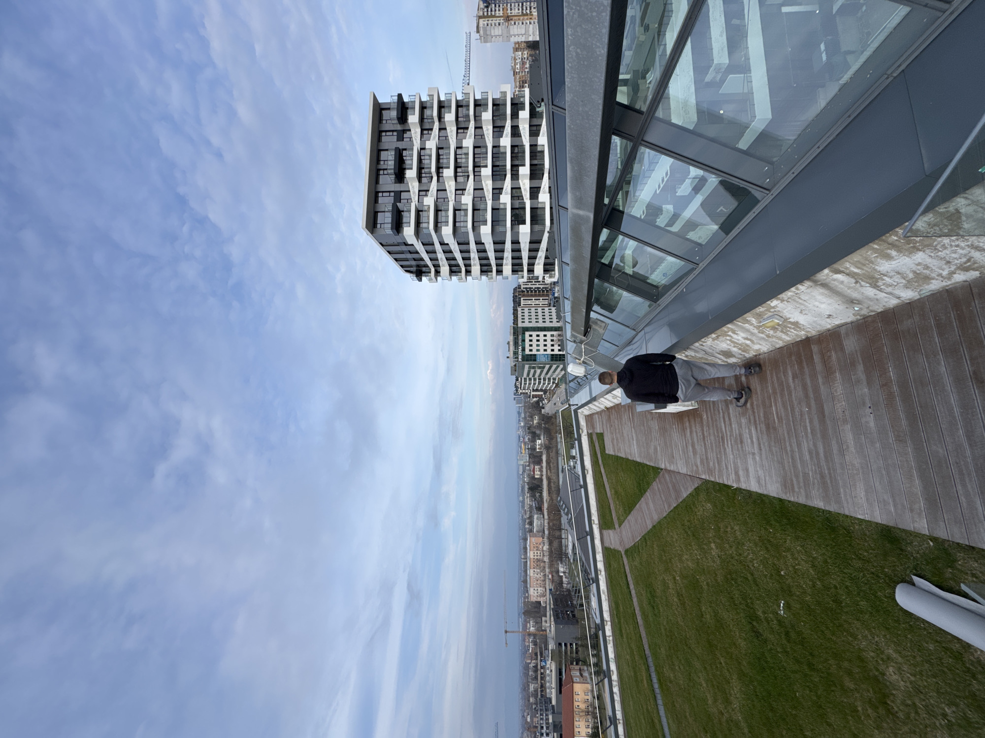 The image captures a perspective from a rooftop in Sofia, Bulgaria. A person stands on a wooden deck with an adjacent green, grassy area. The rooftop architecture includes a modern glass and metal structure. In the background, the city of Sofia unfolds, revealing a mix of older and newer buildings, with at least one construction crane visible. The sky is partly cloudy.