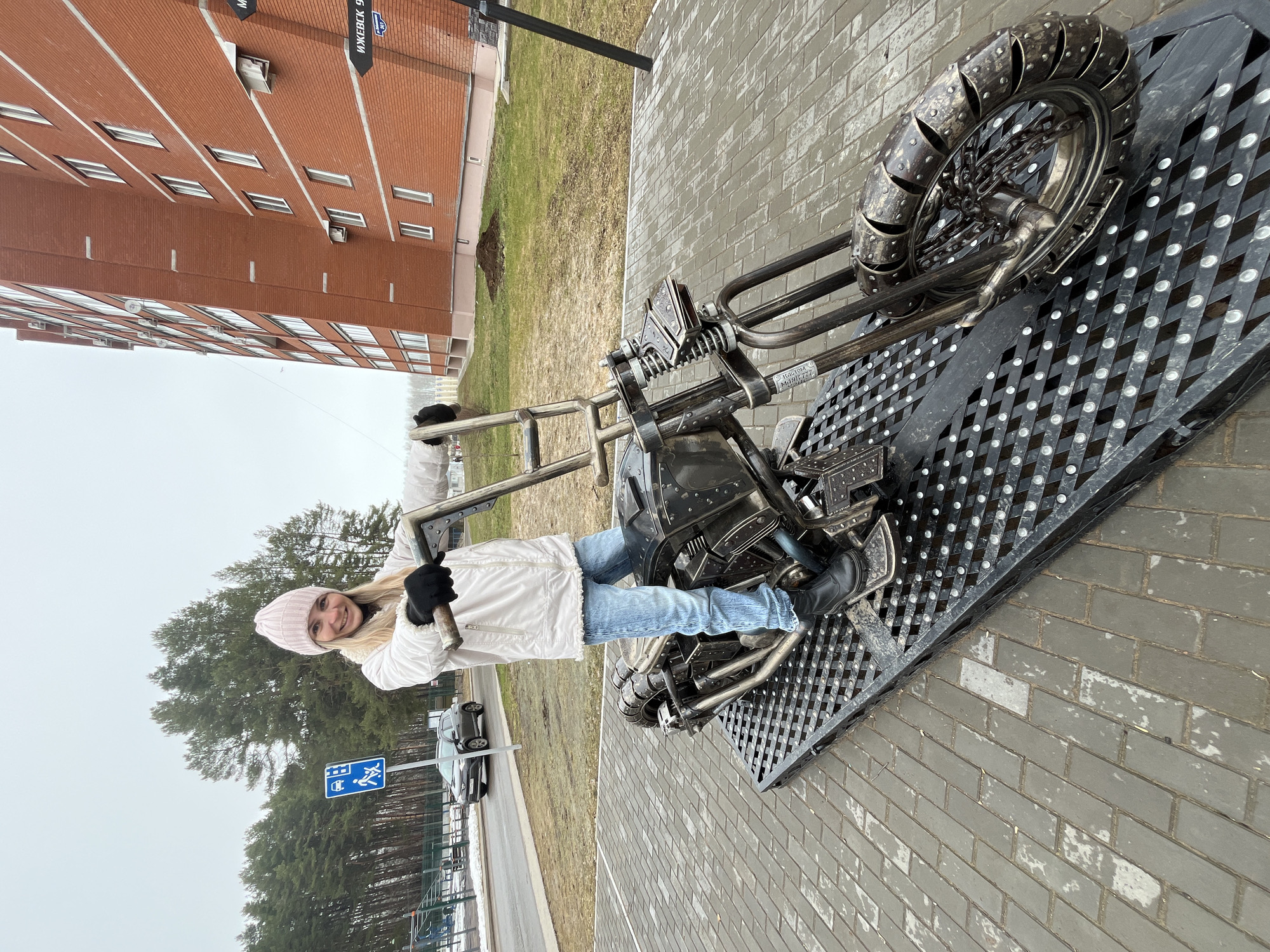 A woman is standing on a metal sculpture of a motorcycle. She is wearing a white jacket, blue jeans, black gloves, and a pink hat. The sculpture is placed on a metal grate on a brick walkway. In the background, there is a tall brick building, a grassy area, trees, and a road with a car. A sign with blue markings on it is nearby. This scene appears to be in Chaikovsky, Russia, according to a street sign that reads "ИЖЕВСК 9-Й МКР-Н".