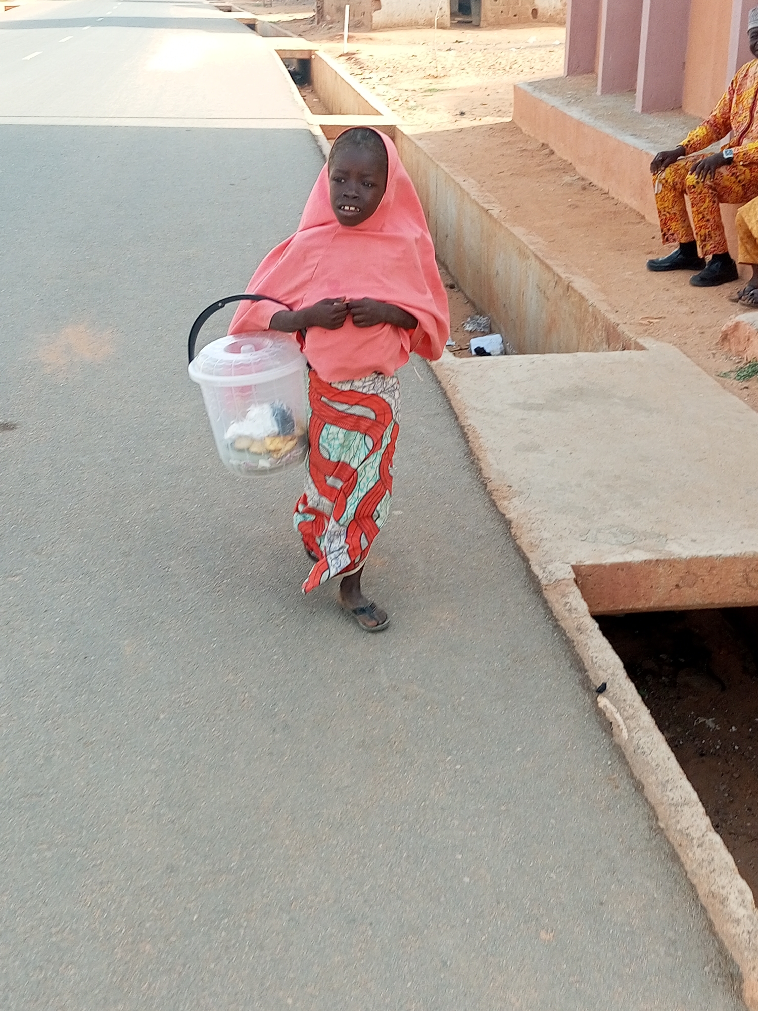 A young girl stands on a paved road in Tempure, Nigeria. She wears a pink headscarf and a patterned wrap-around skirt. She carries a translucent plastic bucket with a black handle, which contains miscellaneous items. In the background, a man is seated on a ledge. A concrete drainage ditch runs along the side of the road. The road stretches towards the horizon.