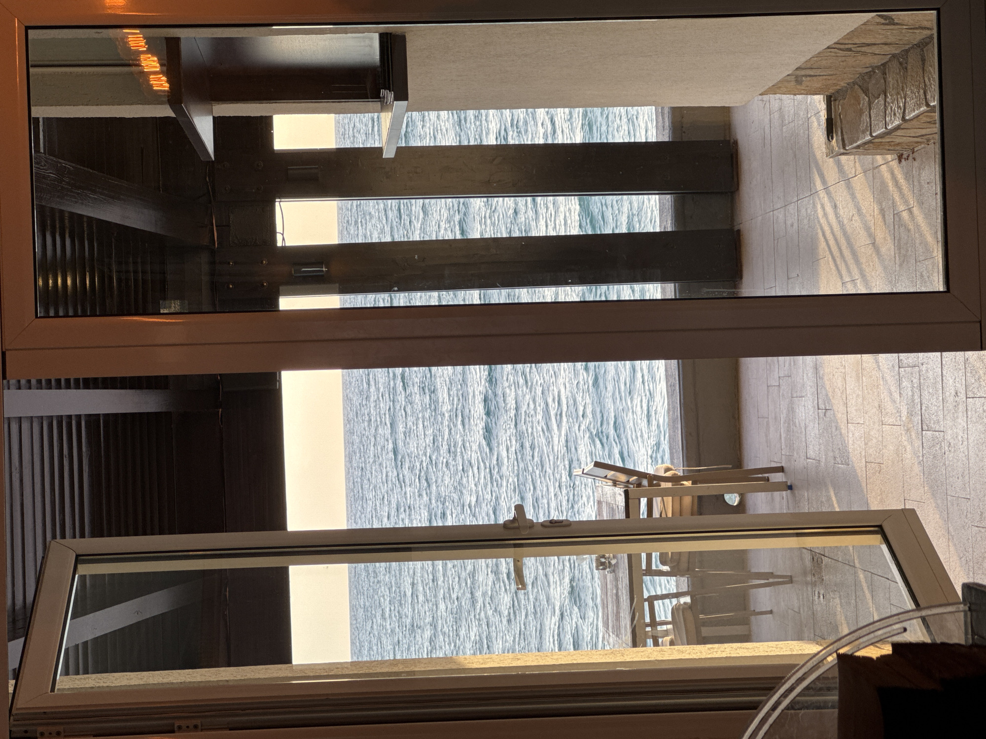 The image shows a view of the sea from what appears to be a building in Bar, Montenegro. The view is seen through a partially opened window with a white frame. In the background, there are sun loungers and tables on a terrace overlooking the sea. The sea itself is visible, with waves reflecting sunlight. Parts of the interior of the building are visible at the top left corner and bottom left corner of the image, suggesting a restaurant or lounge area.