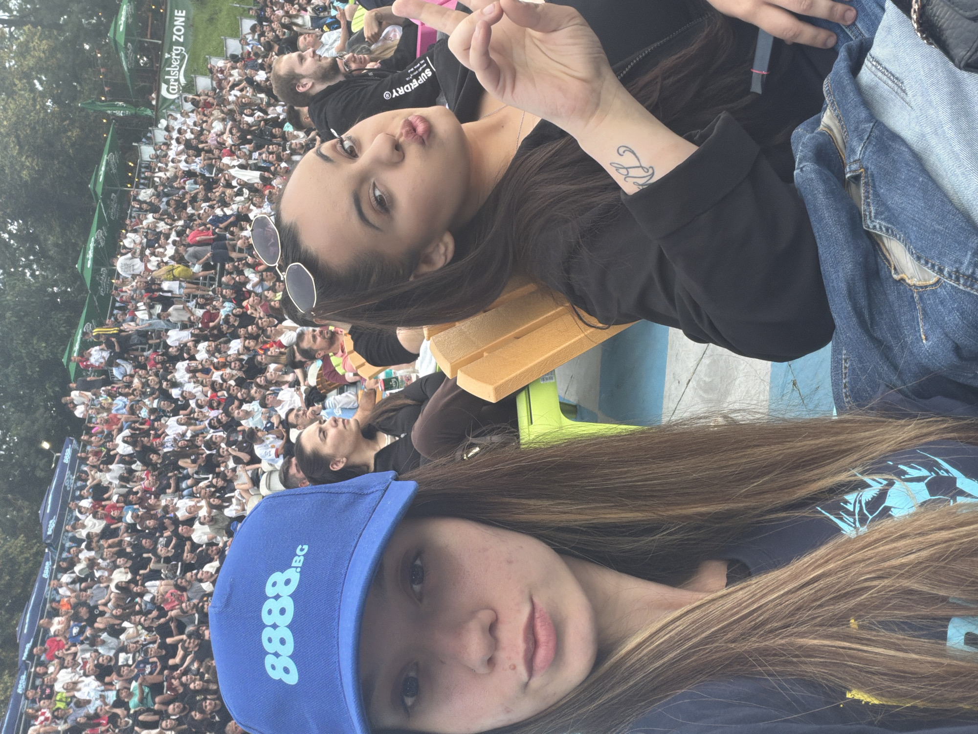 This is a vertical, candid selfie taken outdoors at a crowded event, likely a concert or festival, in Sofia, Bulgaria. Two young women are the primary subjects. The woman in the foreground, wearing a blue baseball cap with light blue text "8888.BG", is looking towards the camera with a neutral expression. Her long, light brown hair is visible. Behind her, a second woman, also with dark brown hair, is leaning back and looking upwards with a playful expression, puckering her lips. She has sunglasses perched on her head and a tattoo on her left forearm.
The background is filled with a large crowd of people, many of whom are looking in the same direction, suggesting they are watching a performance. Various shades of clothing are visible, indicating a casual outdoor event. Green tents with "Carlsberg Zone" text are visible in the upper left portion of the image, providing a clue to the event's sponsorship and nature. The lighting suggests it is daytime, possibly late afternoon or early evening. The overall mood appears to be relaxed and fun.