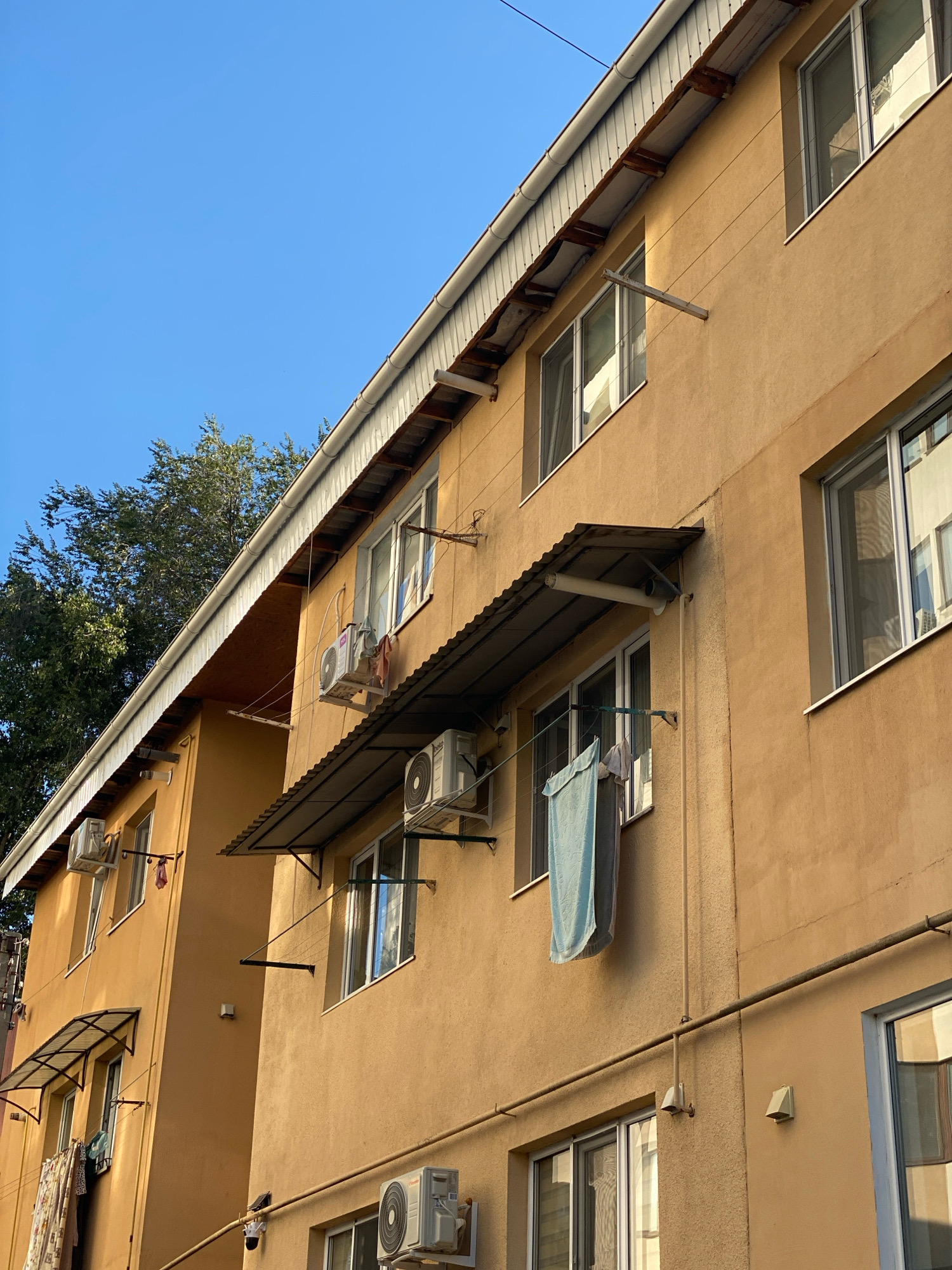 This image shows the exterior of a residential building in Chișinău, Moldova. The building has a light orange stucco facade and is several stories high, with multiple windows and balconies visible. Air conditioning units are mounted on the exterior walls below several of the windows. Clothes are hanging on a clothesline strung between two brackets on one of the balconies, with a light blue towel prominently displayed. The building's roof has a metal overhang supported by wooden beams. A mature tree with dense green foliage is visible to the left of the building, and the sky is clear and blue. The scene appears to be captured during daylight hours, likely in the morning or late afternoon, given the angle of the sunlight casting shadows.