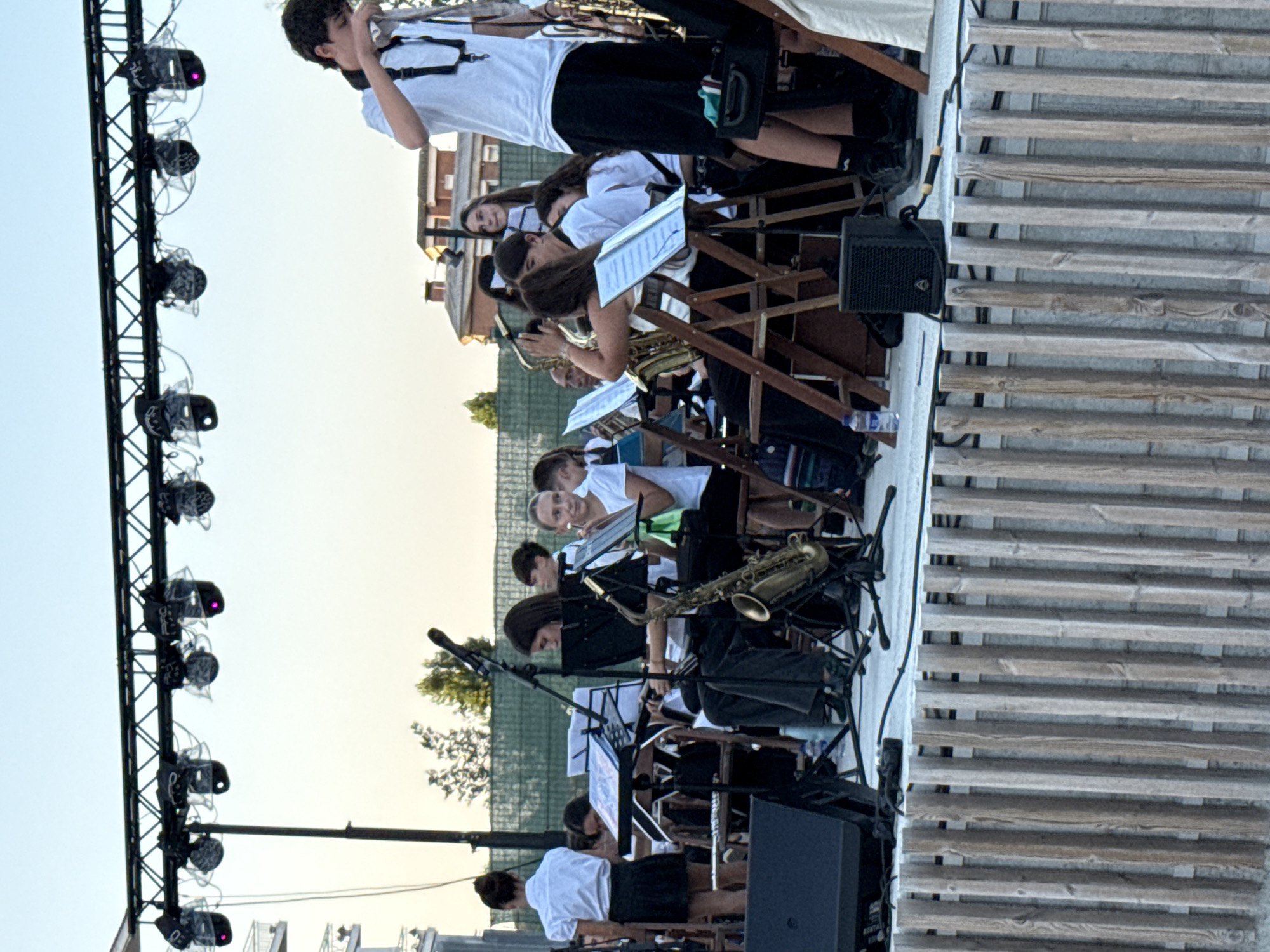 The image depicts an outdoor performance by a musical ensemble, likely a band or orchestra, on a raised wooden stage. The stage is framed by a rustic wooden fence in the foreground. In the background, a clear blue sky is visible, with some buildings and trees suggesting an urban or suburban setting. A lighting rig with multiple spotlights is suspended overhead, indicating a professional setup for a performance.
The ensemble appears to consist primarily of young people, both male and female, dressed in white tops and dark bottoms, likely a uniform. They are seated on chairs, positioned around music stands, and are engaged in playing various instruments. Visible instruments include saxophones, a clarinet, and possibly flutes. Some individuals are actively playing their instruments, while others are looking at their sheet music. One individual in the foreground, on the right side of the image, is standing and holding a saxophone, seemingly in the process of playing or preparing to play.
The overall atmosphere suggests a lively event, possibly a concert or festival. The presence of musical instruments, the stage, lighting, and the attentive audience (implied by the performance setting) all contribute to this impression. The city of Blanes, Spain, is known for its cultural events and festivals, and this scene could plausibly be part of such an occasion.