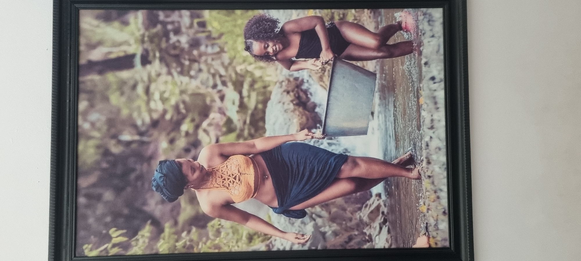 A framed photograph hangs on a white wall. The photograph depicts two women in a natural environment that may be a river or stream. The woman in the foreground is wearing a tan top, and a dark-colored wrap. The woman in the background is wearing a black, strapless top and holding a metal container. The background features blurred vegetation and rocks. The frame is black. There is no information provided to determine whether the location is in Lekki, Nigeria.