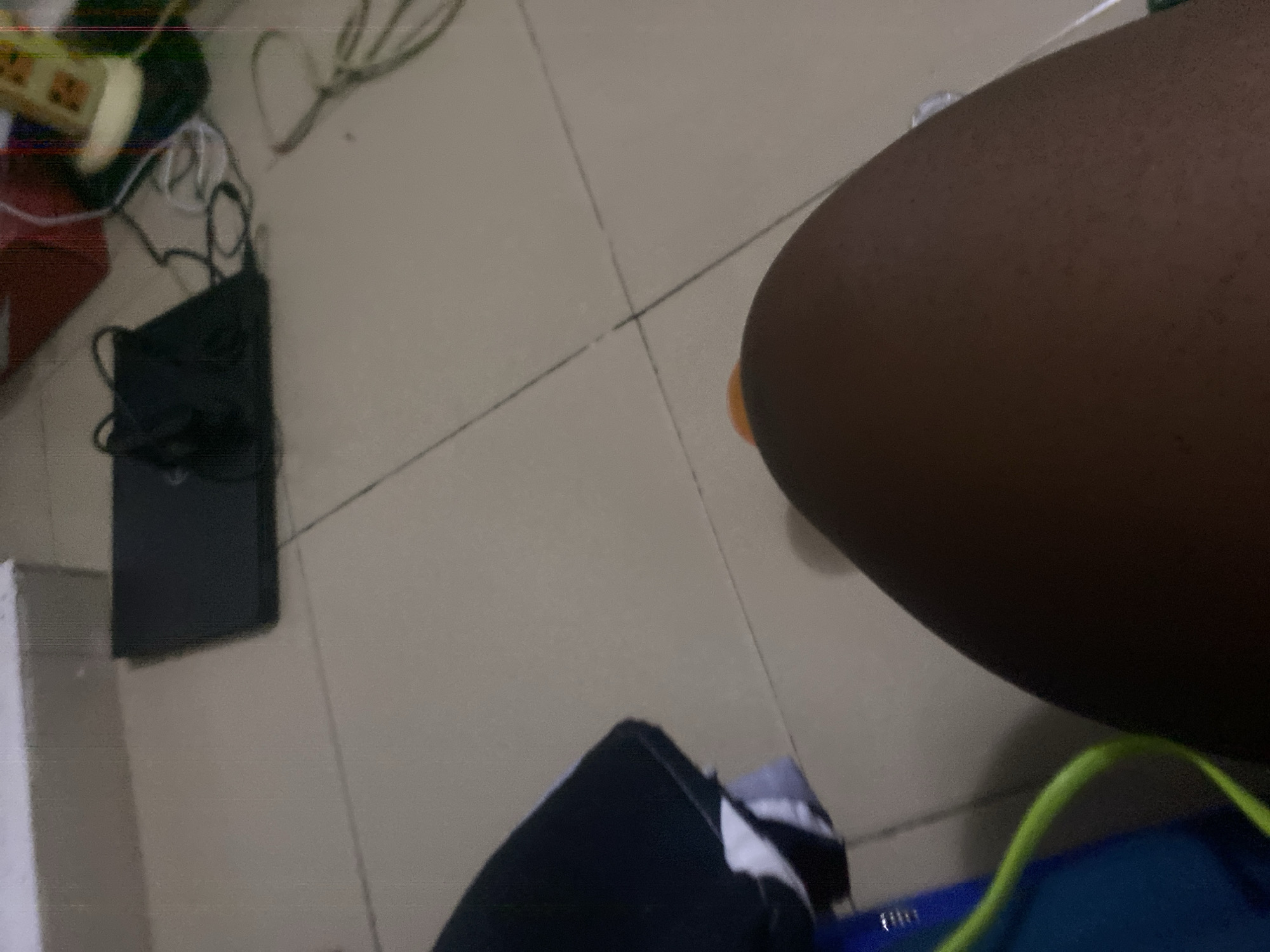 The image depicts a tiled floor in what appears to be an indoor setting in Howayi, Nigeria. The floor tiles are light beige with darker grout lines. In the upper left portion of the image, a white rectangular object, possibly a box or piece of furniture, is visible. To its right, a black laptop is placed on the floor, connected to charging cables. Further to the right, a red box is partially visible. In the foreground, a person's dark-skinned leg is prominently featured, with a portion of their thigh and knee visible. A bright green cable snakes across the lower left corner of the image, connected to a blue object. The overall scene suggests a domestic or casual environment, possibly a living room or bedroom.